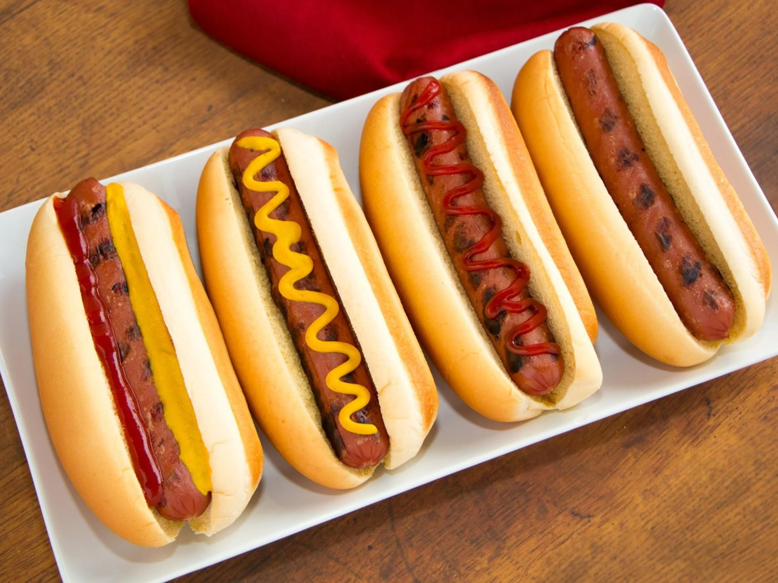 Four hot dogs in buns on a white rectangular plate, each topped with ketchup, mustard, or both, on a wooden table with a red napkin in the background.