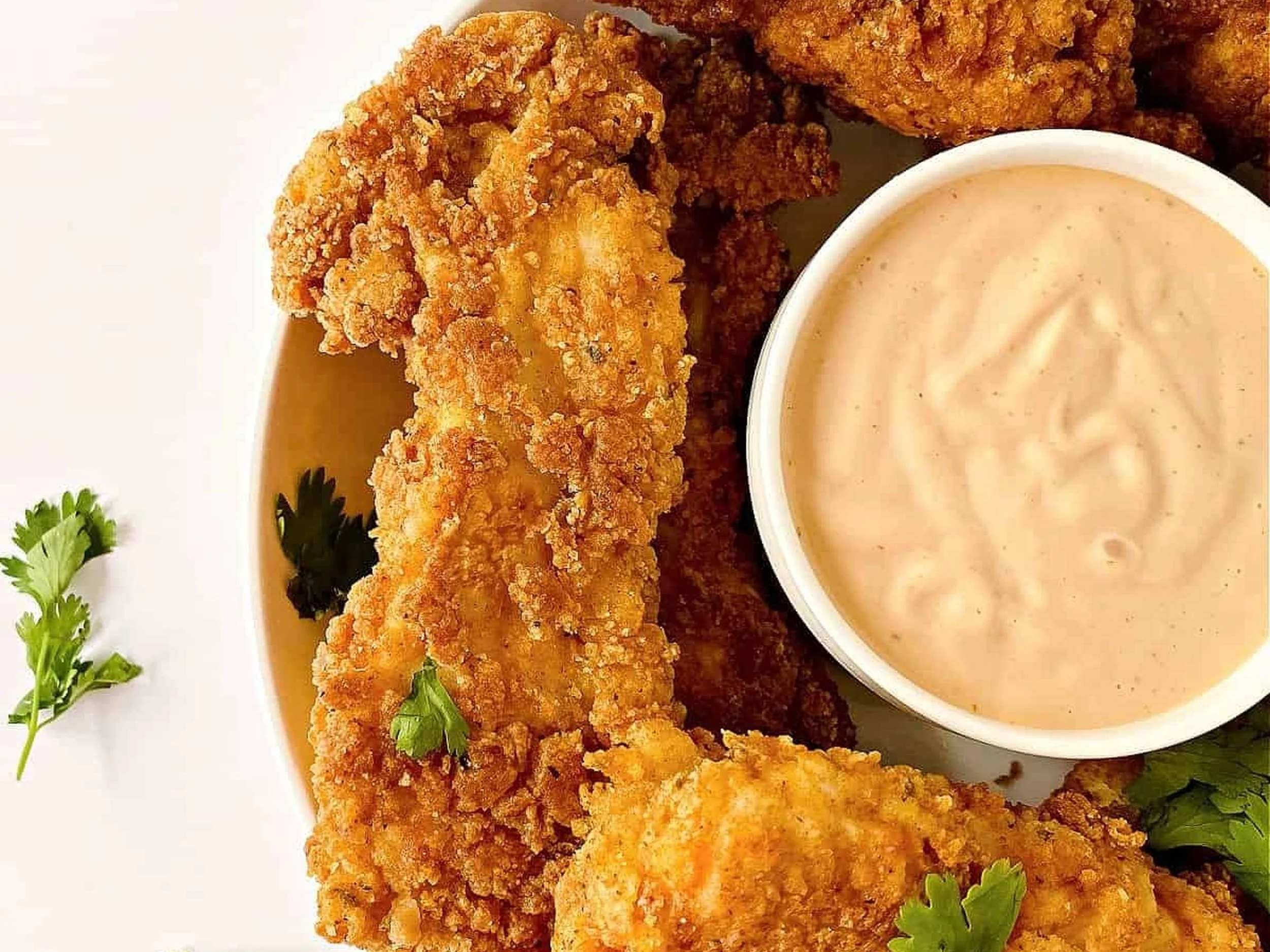 Fried chicken tenders with dipping sauce on a white plate, garnished with parsley and cilantro.