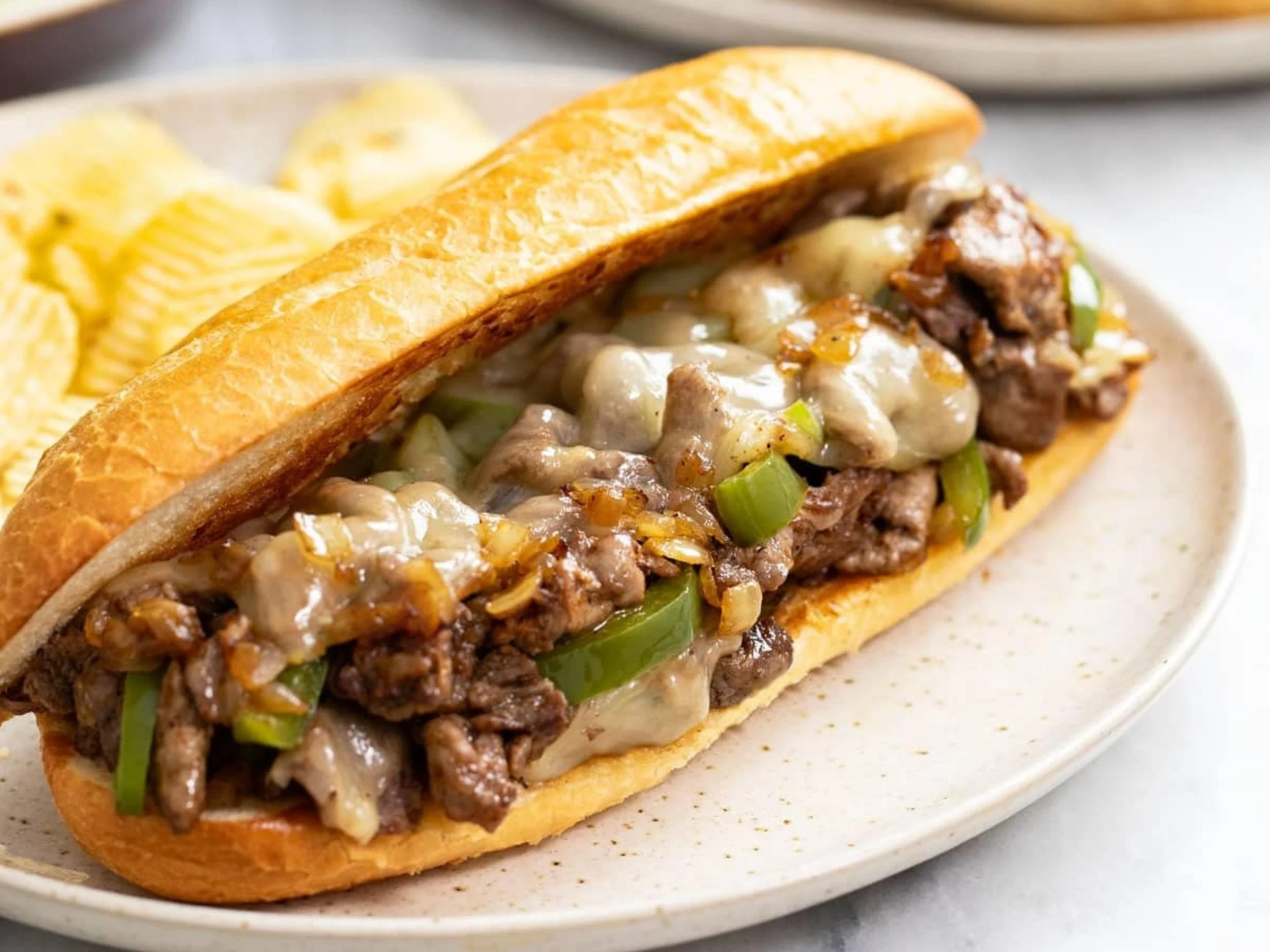Close-up of a beef sandwich with melted cheese, green bell peppers, and onions on a toasted hoagie roll, served on a white plate.
