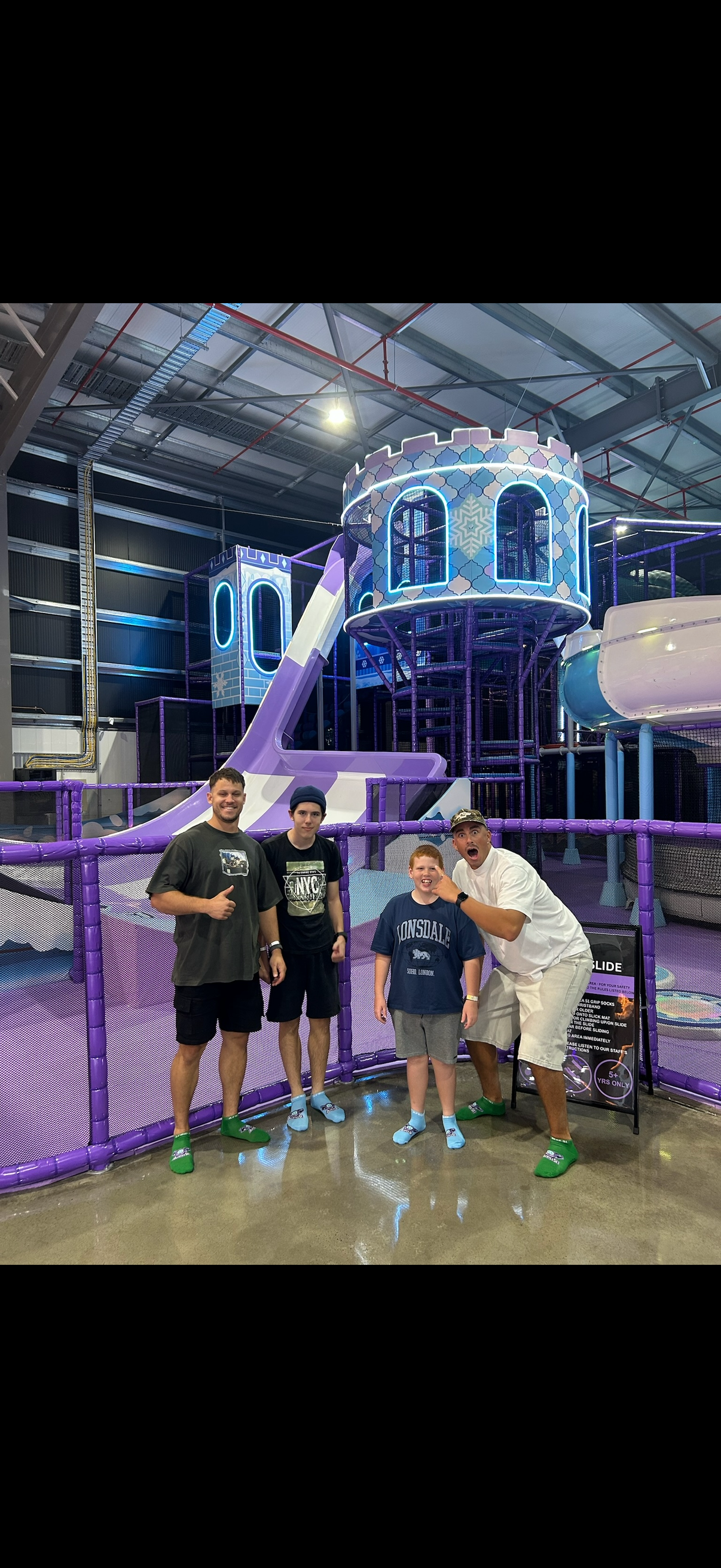 Group of four boys and one man standing in front of an indoor playground with purple and blue slides and structures, smiling and posing for the camera.