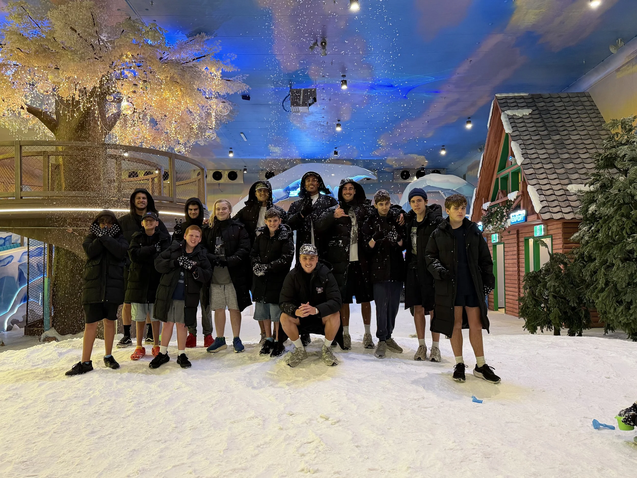 Group of young people standing together inside an indoor snow attraction with winter-themed decor, artificial snow on the ground, and illuminated snow-covered trees and buildings in the background.
