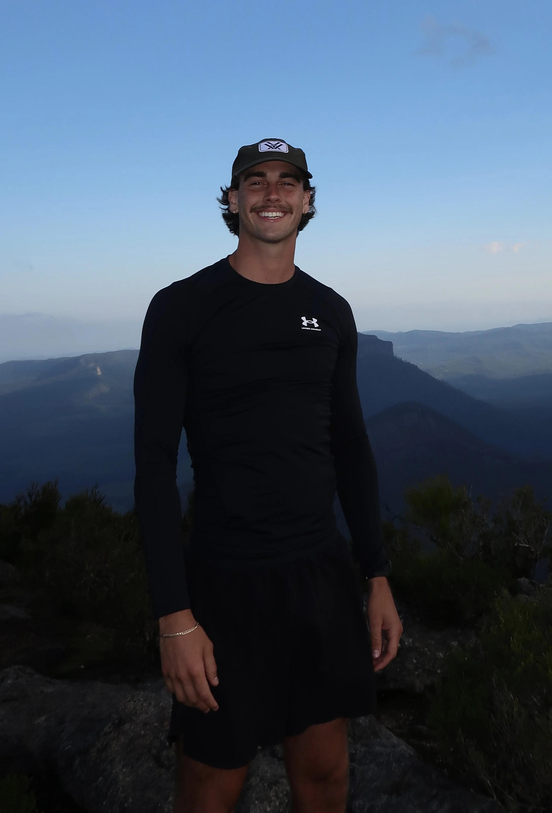 A man standing outdoors on a mountain with a scenic background of hills and sky, smiling at the camera, wearing a black Under Armour shirt and a black cap.