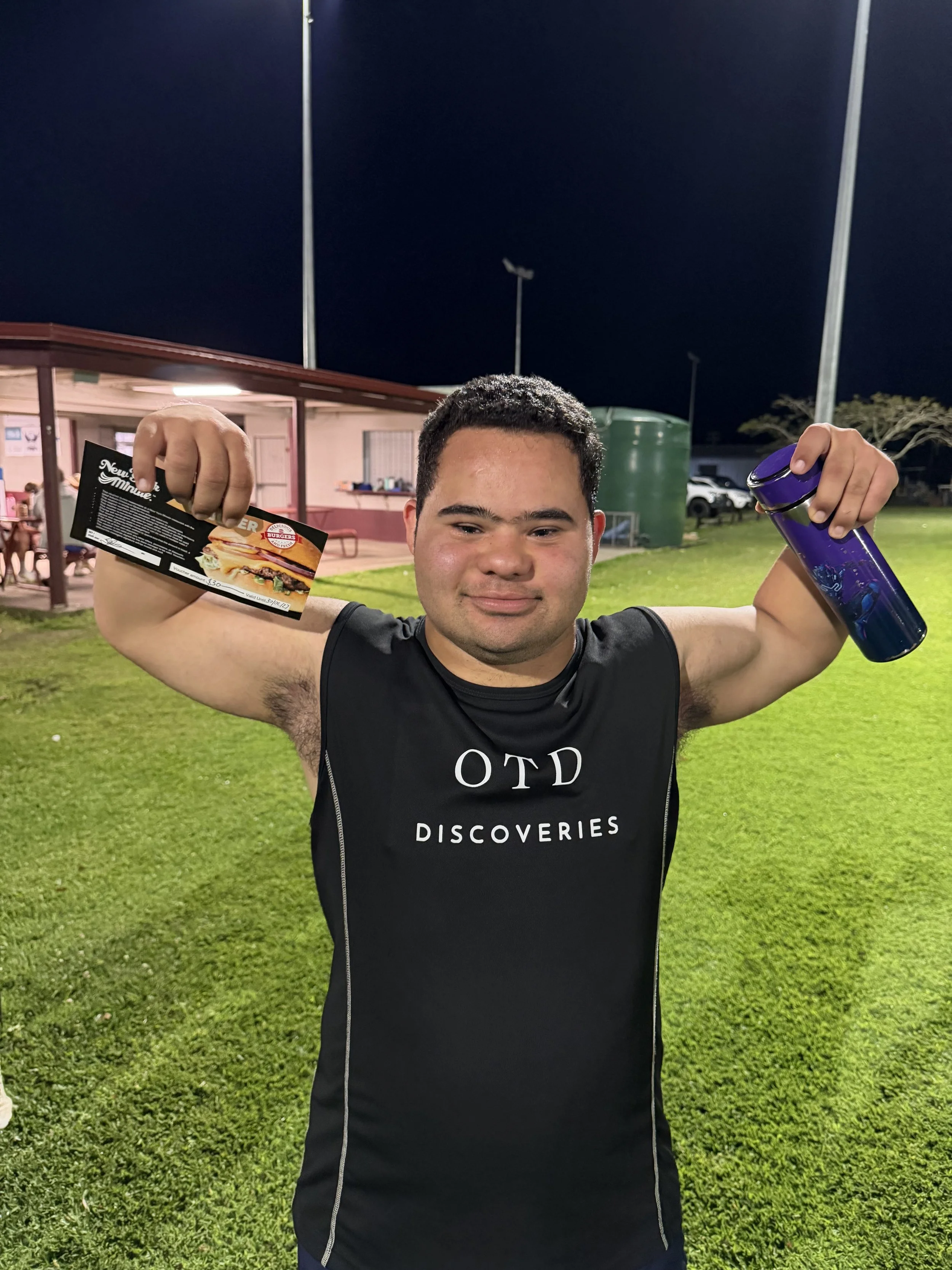 A young man with short dark hair, wearing a black athletic shirt that says 'OTD Discoveries,' stands on a grassy field at night, smiling and holding a promotional flyer in one hand and a purple water bottle in the other.