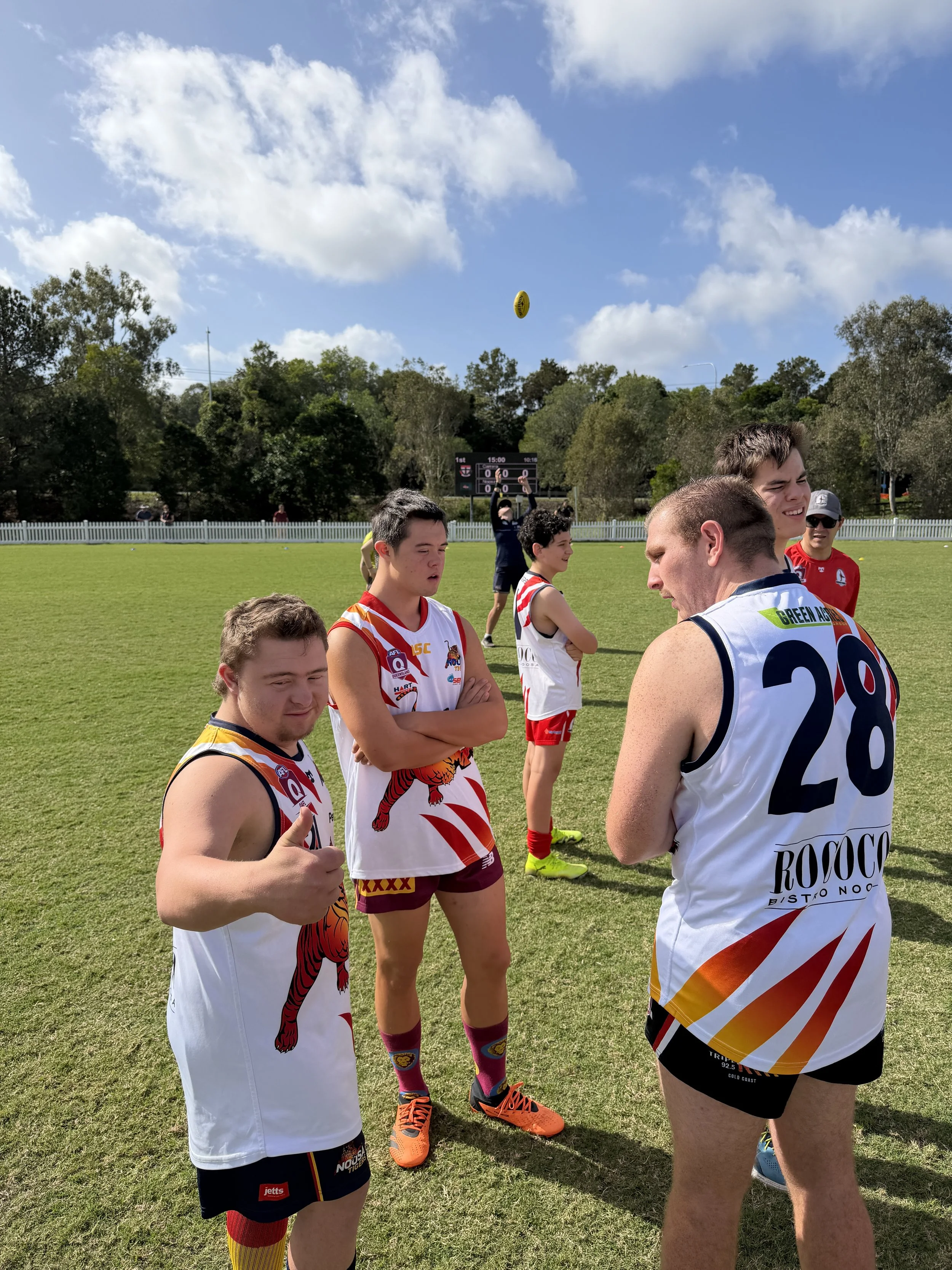 Australian rules football players in colorful uniforms standing on a grassy field, some with arms crossed, during a match on a sunny day with an electronic scoreboard and trees in the background.