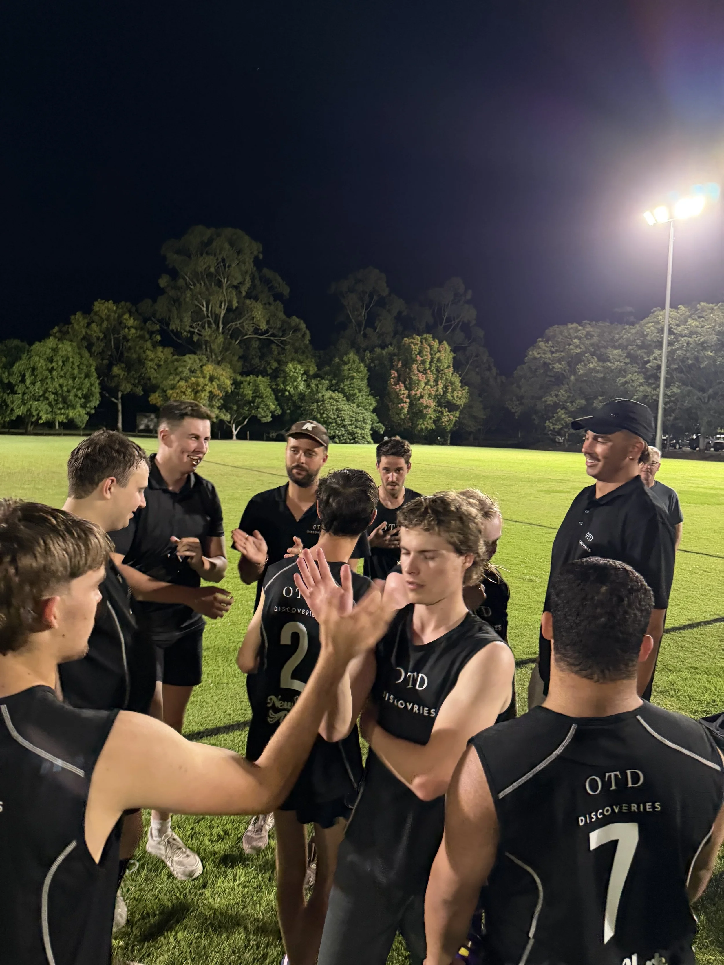 A group of young men wearing black sports jerseys in a huddle on a well-lit outdoor field at night, with trees in the background.