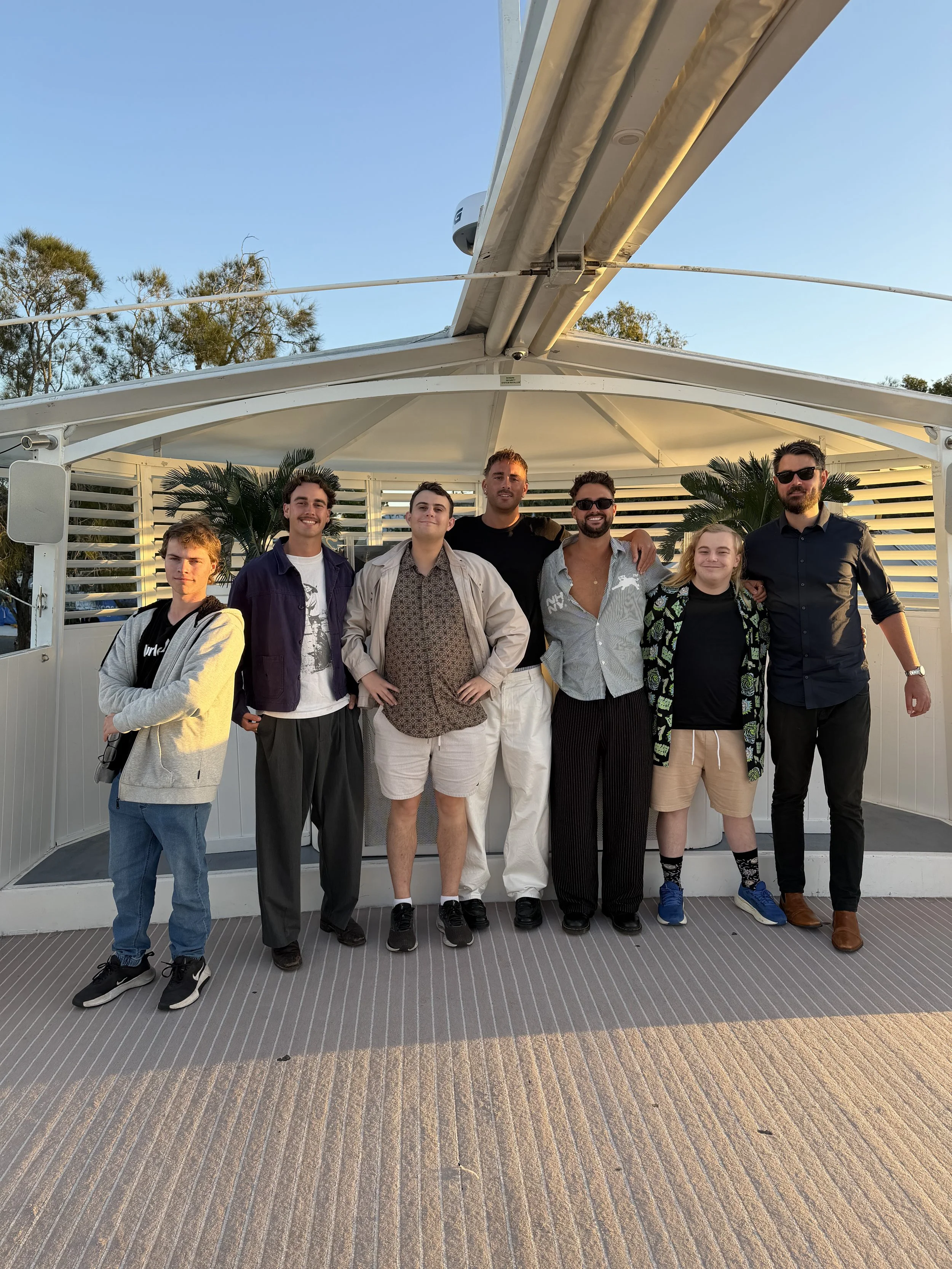 A group of eight young adults standing on a boat deck, smiling and posing for the photo, with some wearing sunglasses, and trees visible in the background.