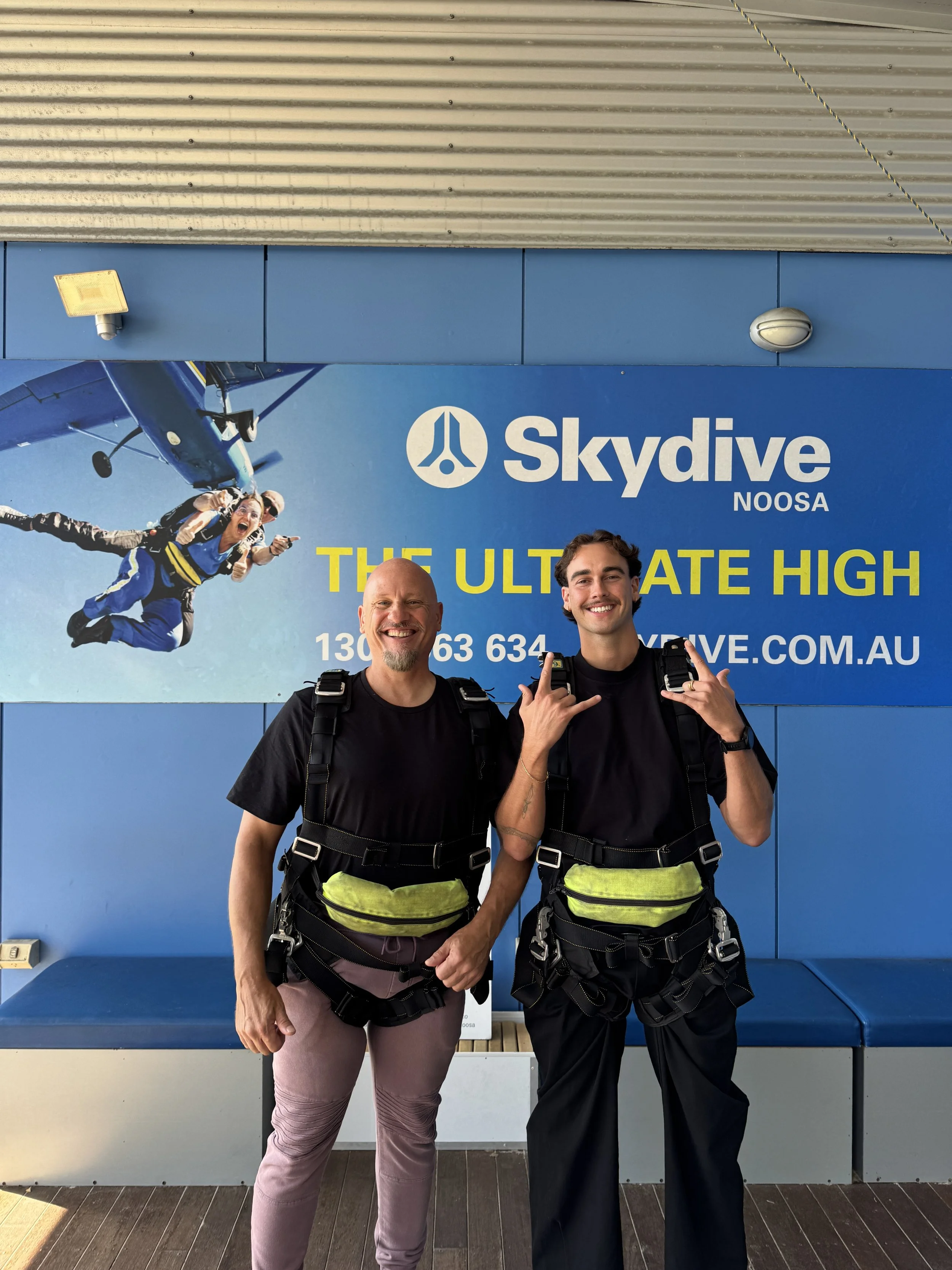 Two men standing in front of a blue skydiving advertisement, both wearing skydiving gear and smiling at the camera.