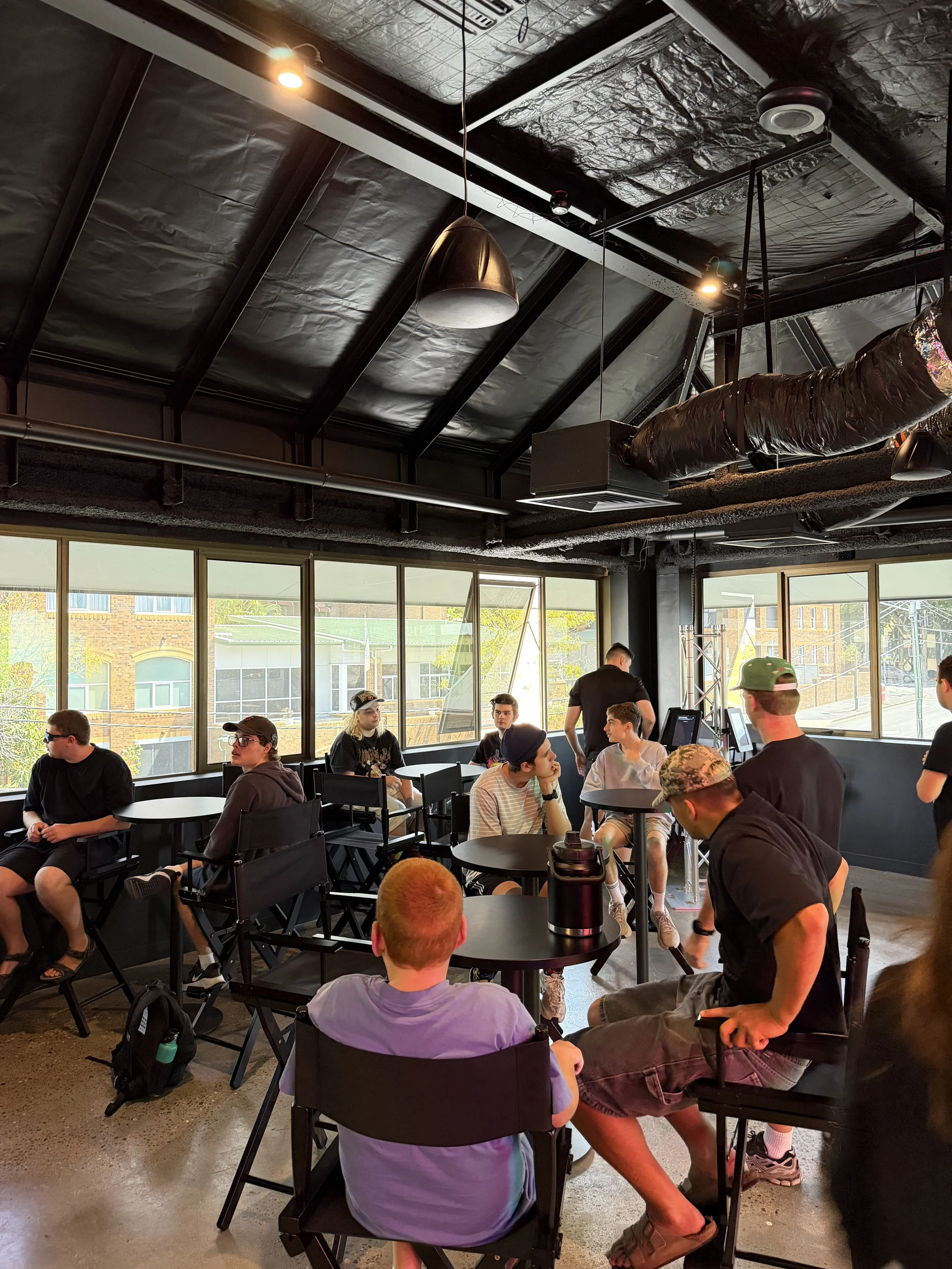 A group of young people sitting and standing around in a modern indoor space with large windows, black tables and chairs, ceiling lights, and visible ceiling ductwork, engaged in conversation.