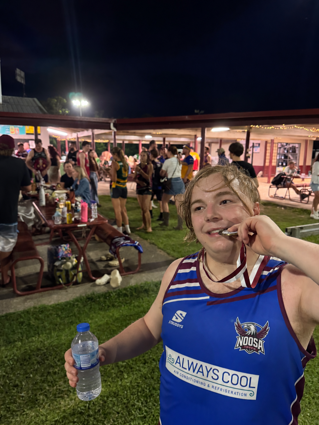 Young man in a blue sports jersey holding a water bottle and a medal, at a nighttime outdoor gathering with people socializing and eating under string lights.