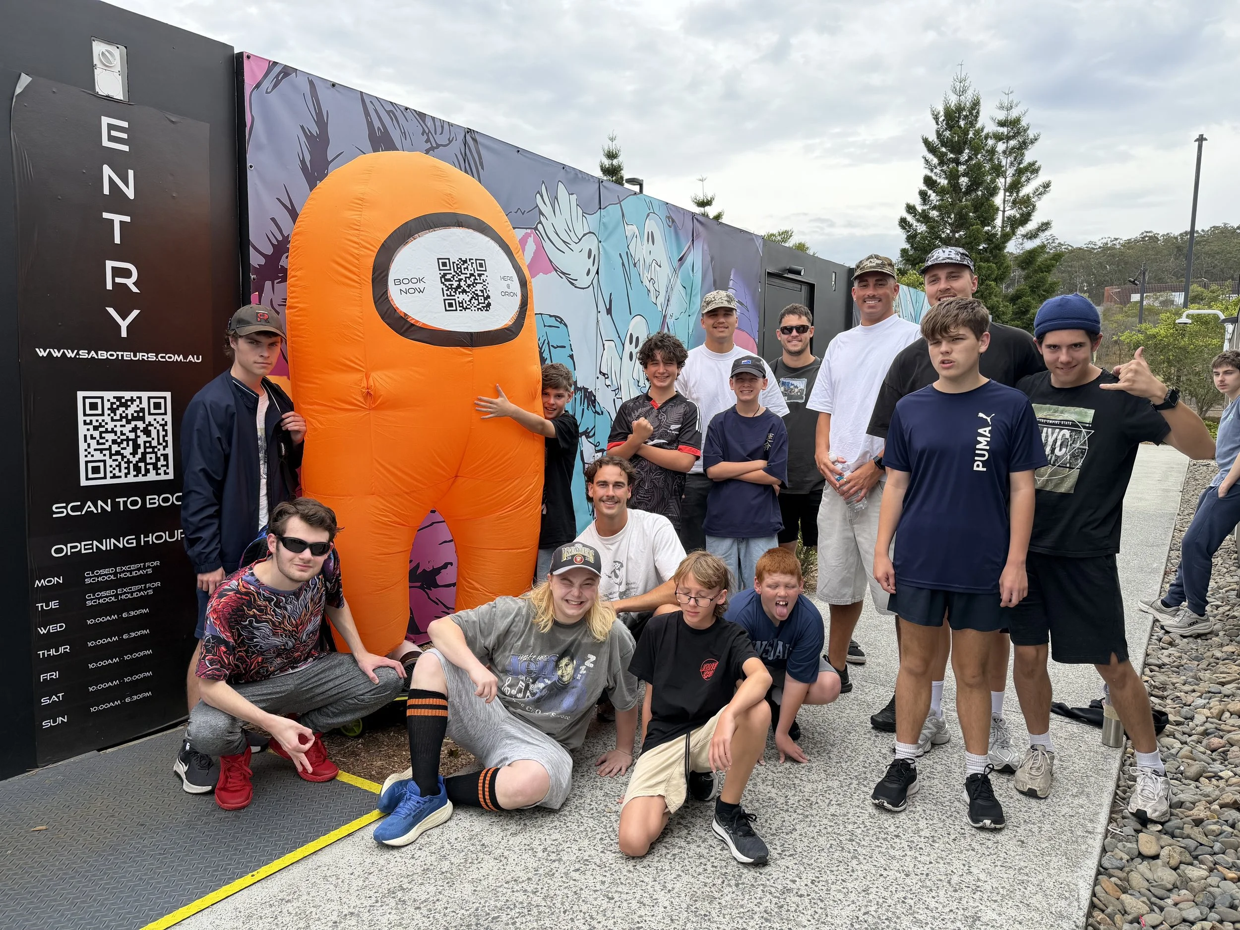 A group of boys and men posing outdoors in front of a colorful illustrated wall and an orange inflatable suit with a QR code. There is a black sign with event details on the left side.