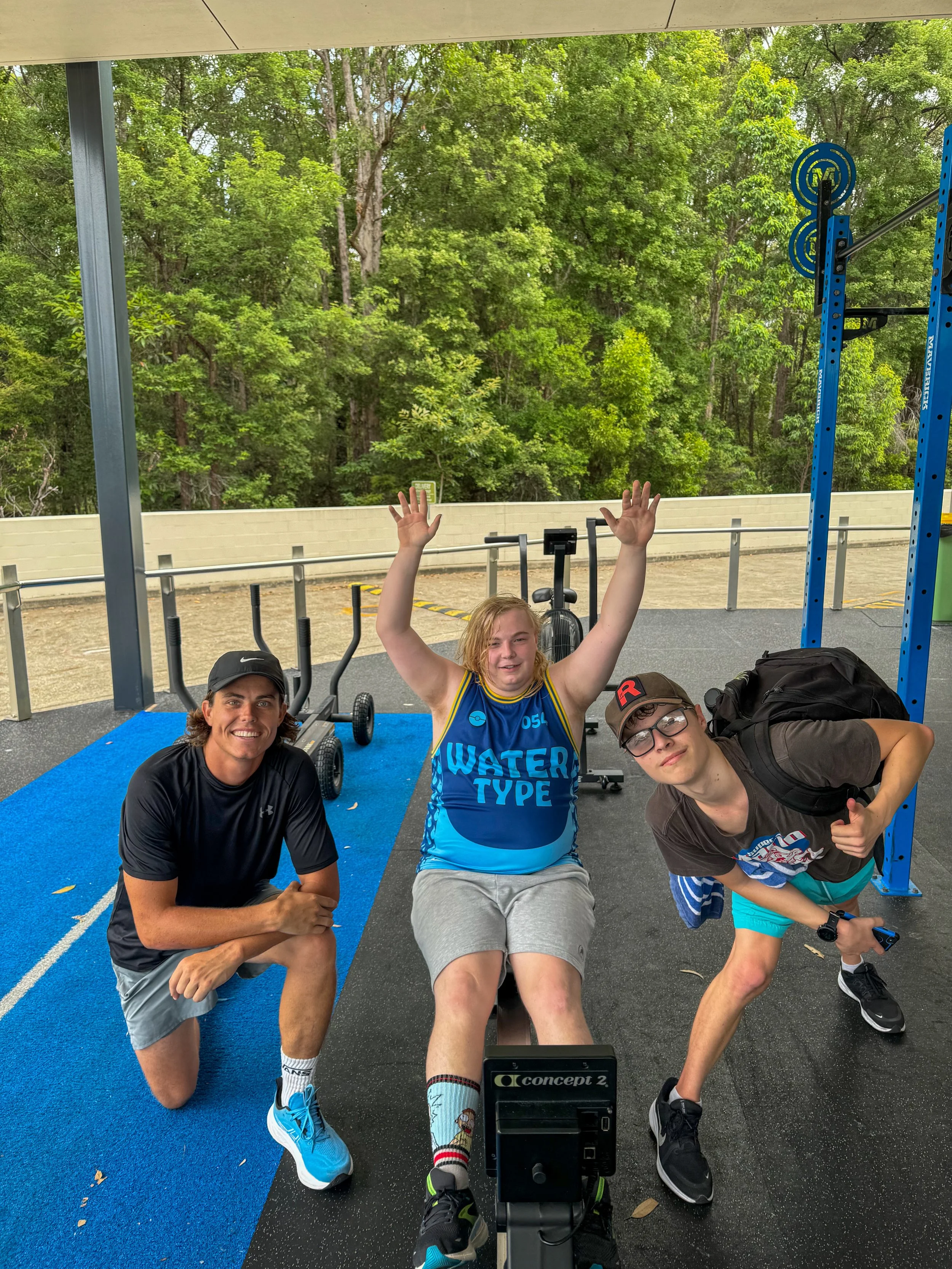 Group of four young people in workout clothes, two kneeling, one sitting in the middle with arms raised, one standing on the right giving a thumbs-up, in an outdoor gym with trees in the background.