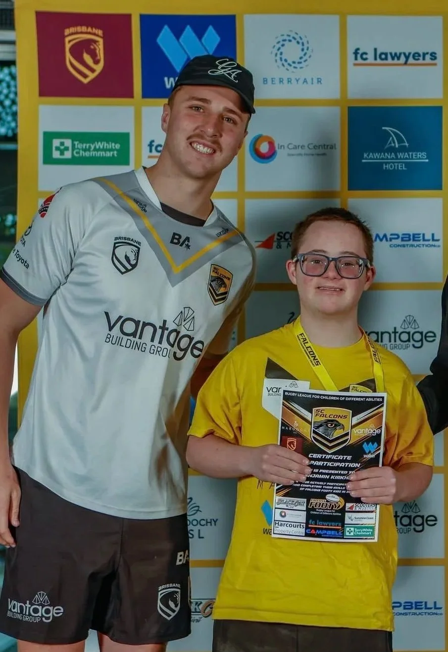 A young man wearing a Brisbane Lions jersey and cap, smiling for a photo with a young boy holding a certificate at a sports event, with a backdrop of various sponsor logos.