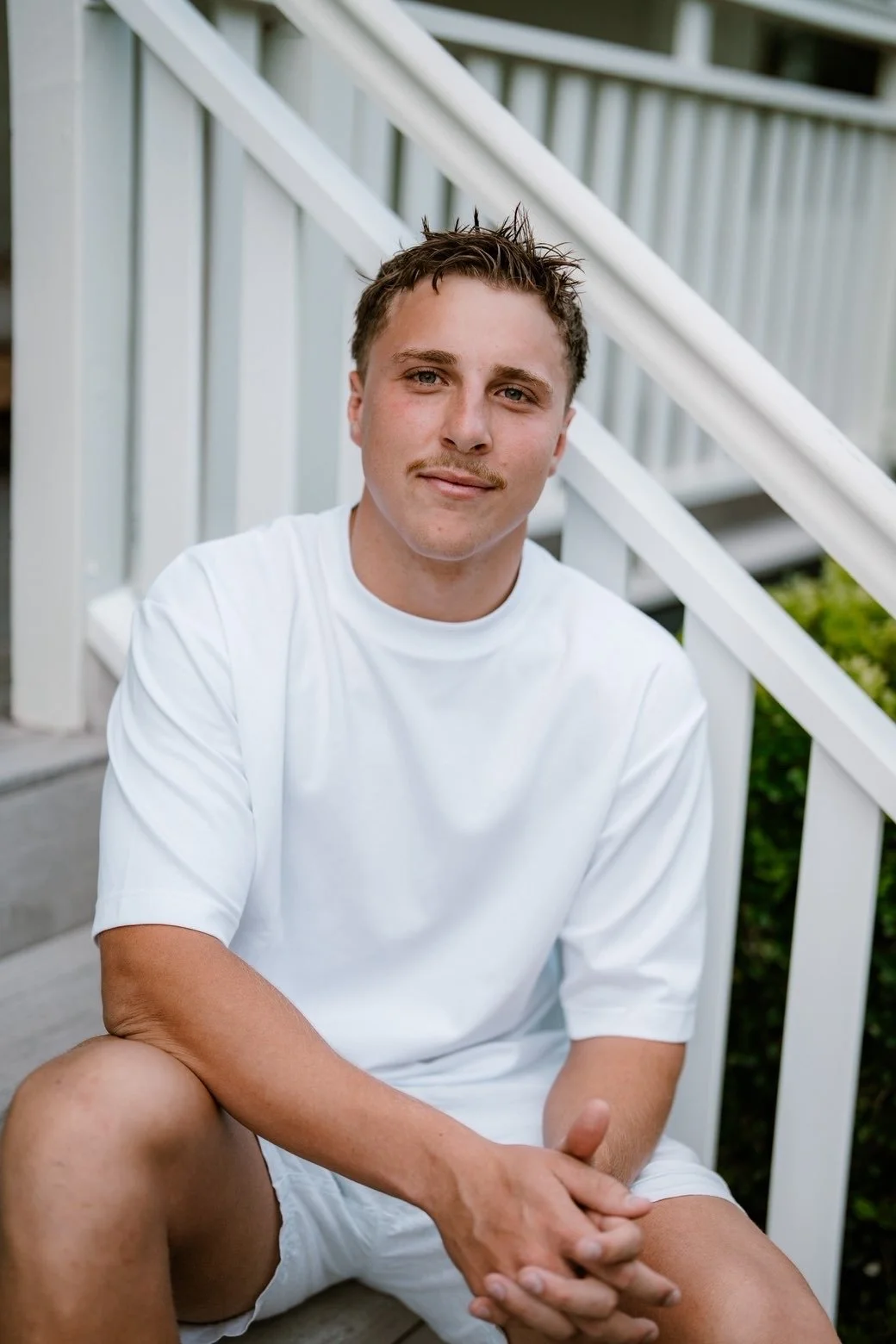 A young man with short brown hair, a light mustache, and fair skin, wearing a white t-shirt, sitting on a concrete surface with legs apart, smiling softly, with white staircase and greenery in the background.