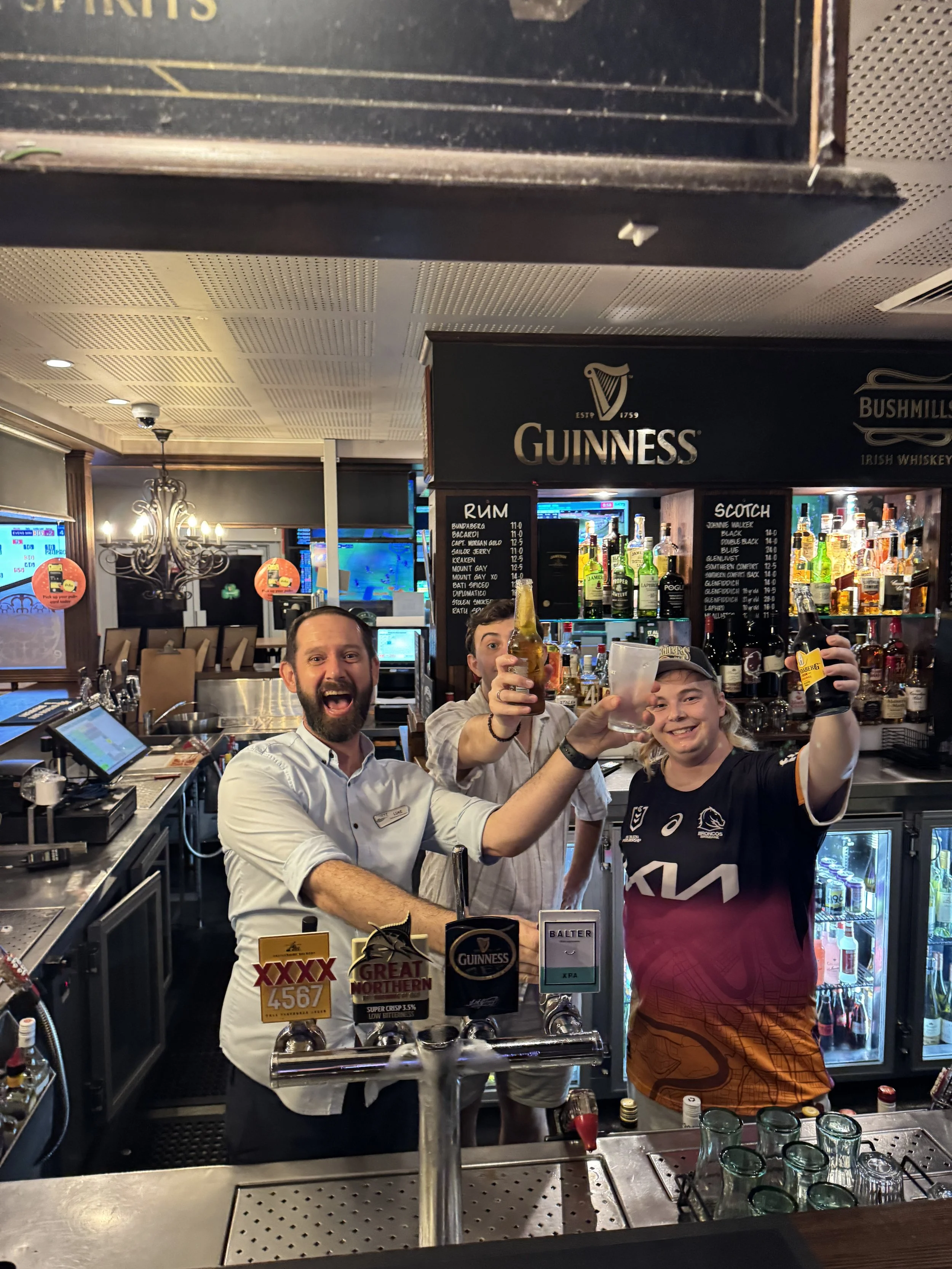 Three smiling bartenders celebrating with drinks in a bar, with Guinness signage and various bottles behind the counter.