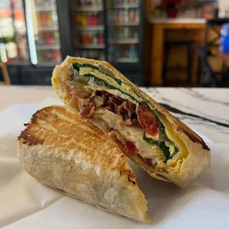 A grilled sandwich cut in half, showing layers of lettuce, tomato, cheese, bacon, and possibly cucumber, on a white surface in a cafe or restaurant setting.