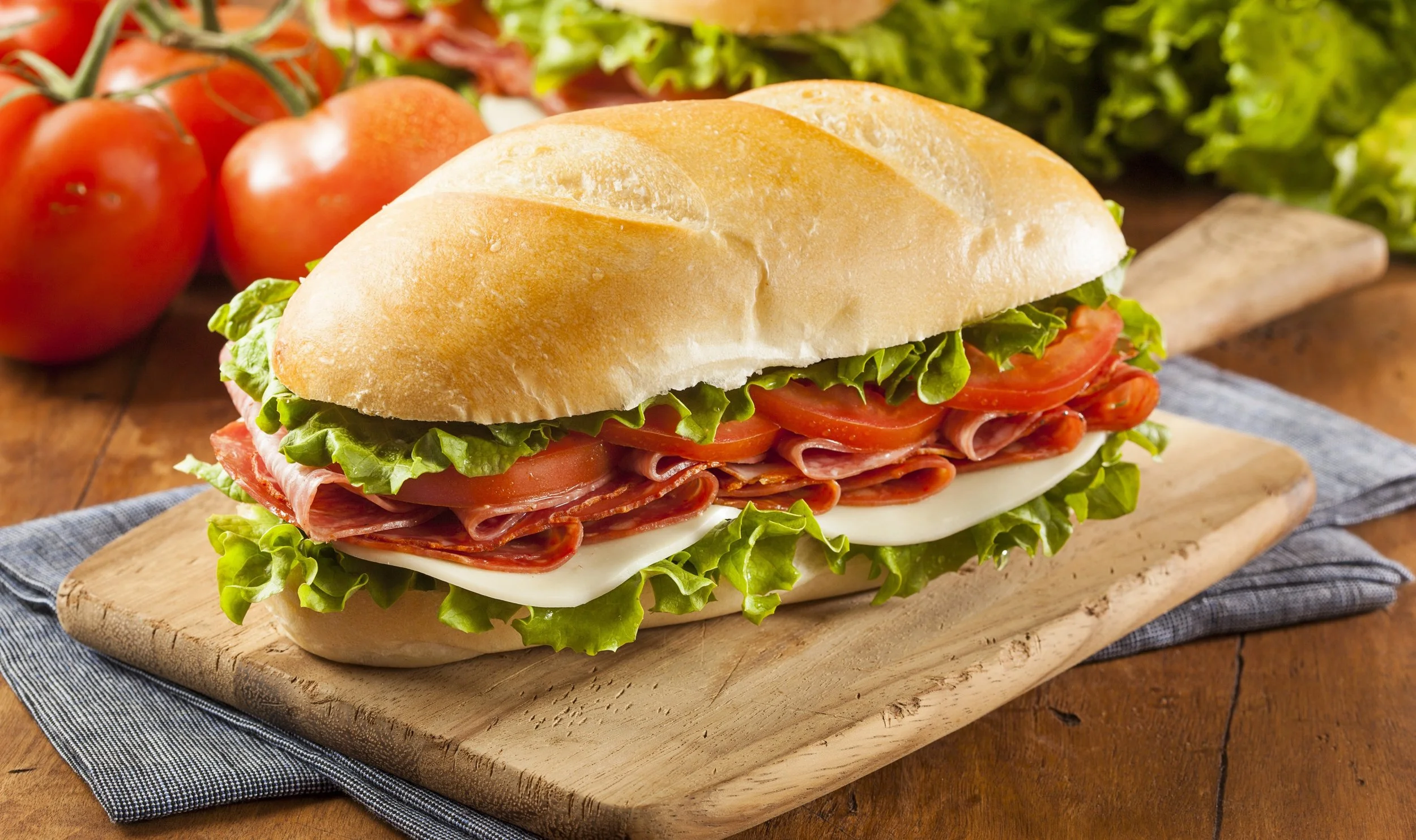 A sandwich with lettuce, tomato, and meat on a bun, placed on a wooden cutting board. Fresh tomatoes and leafy greens are in the background.