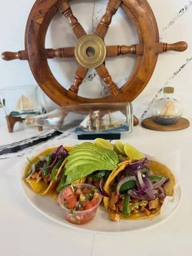 Two tacos with meat, lettuce, tomatoes, and purple onions on a white plate, with a small bowl of salsa in front, on a white table with nautical decor including a ship's wheel and miniature sailboats in the background.