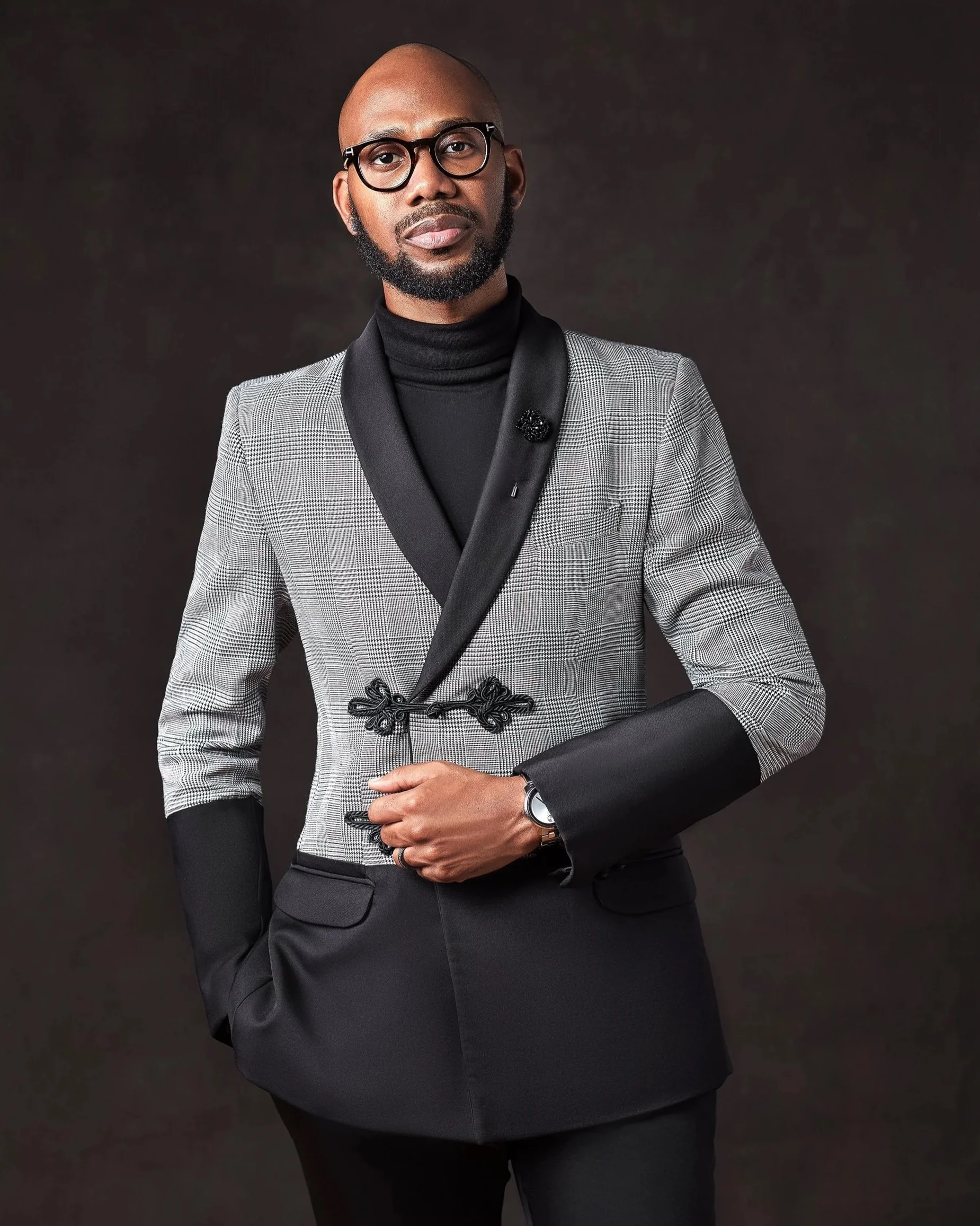A professional African American man wearing glasses, a black turtleneck, and a gray checkered blazer with black lapels, posing against a dark background.