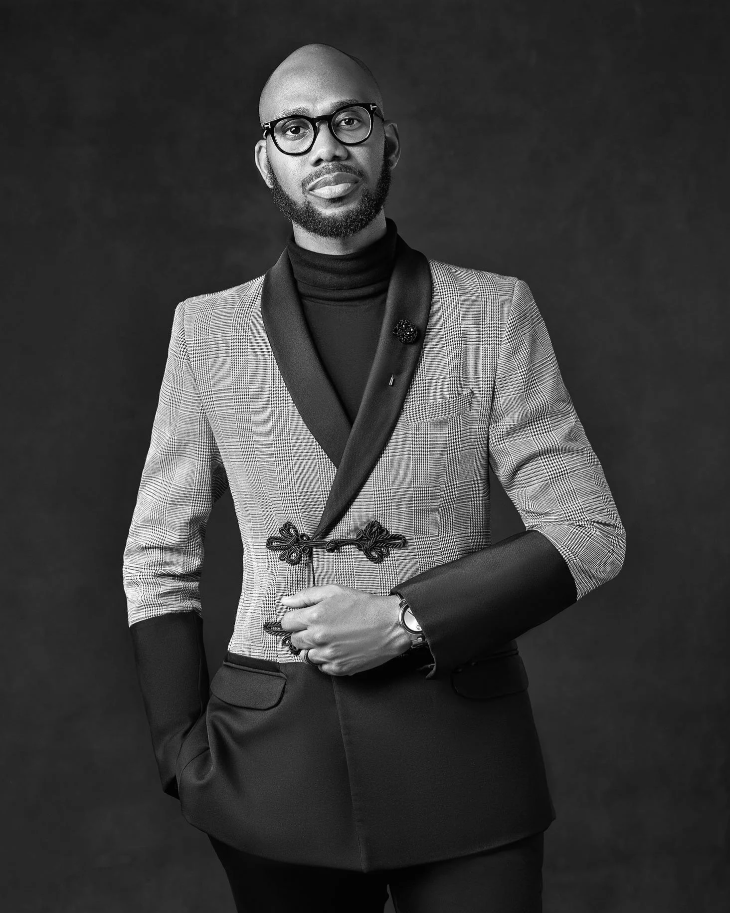 Black and white photo of a well-dressed man wearing glasses and a patterned blazer, standing against a dark background.