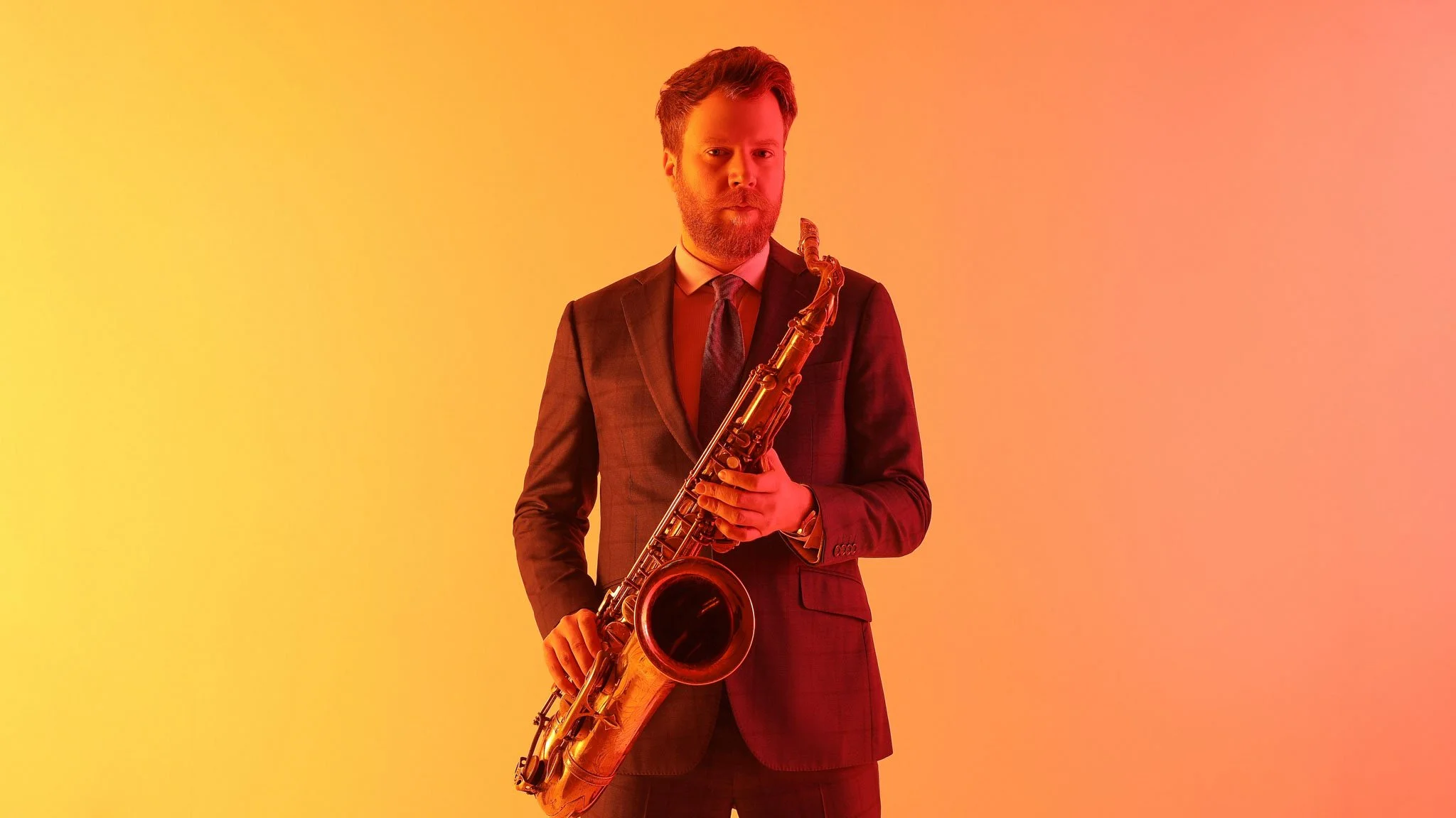 A man with a beard wearing a suit, holding a saxophone, standing against a gradient background of yellow, orange, and pink.