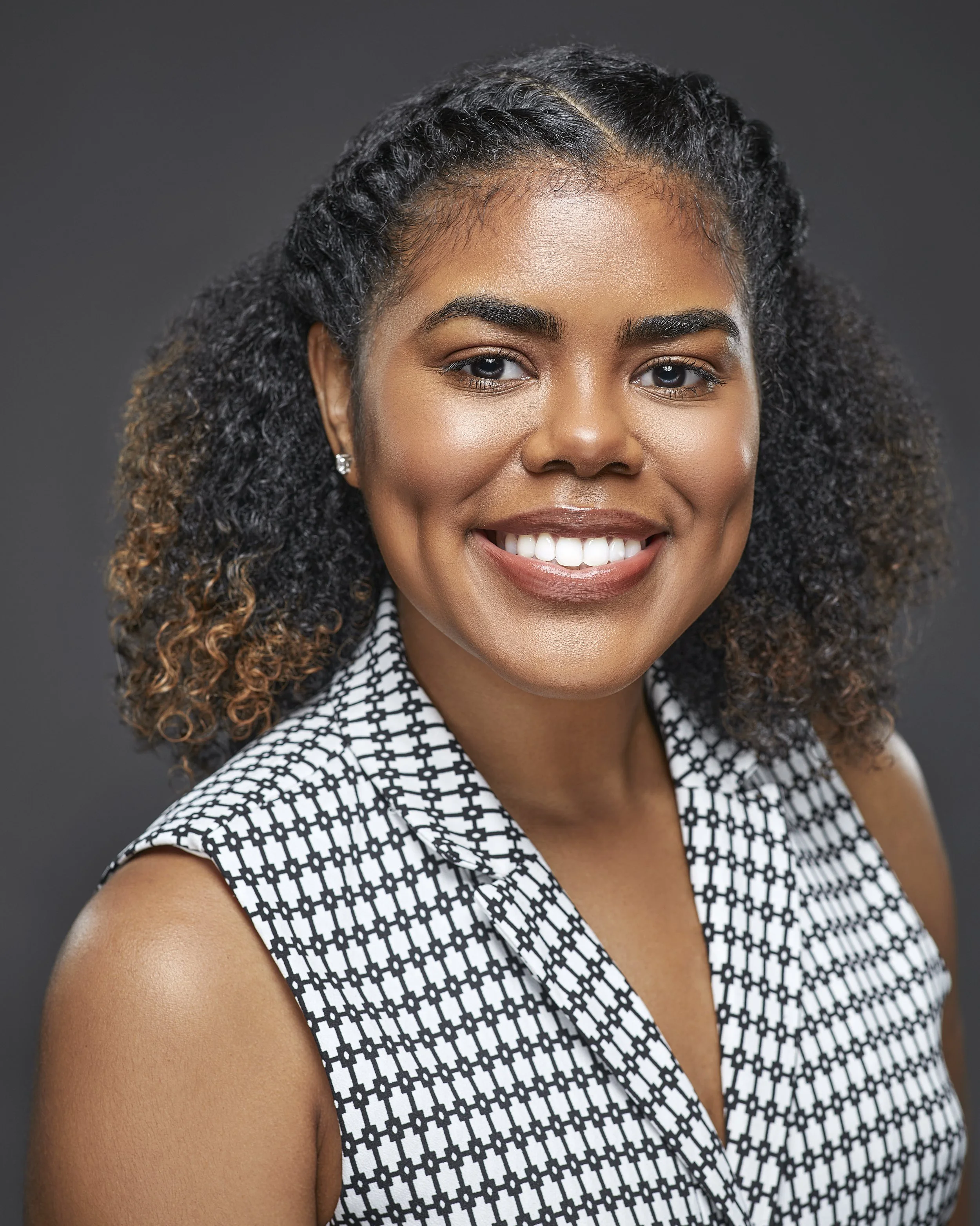A woman with curly hair smiling at the camera, wearing a sleeveless black-and-white patterned top, and silver earrings, against a dark gray background.