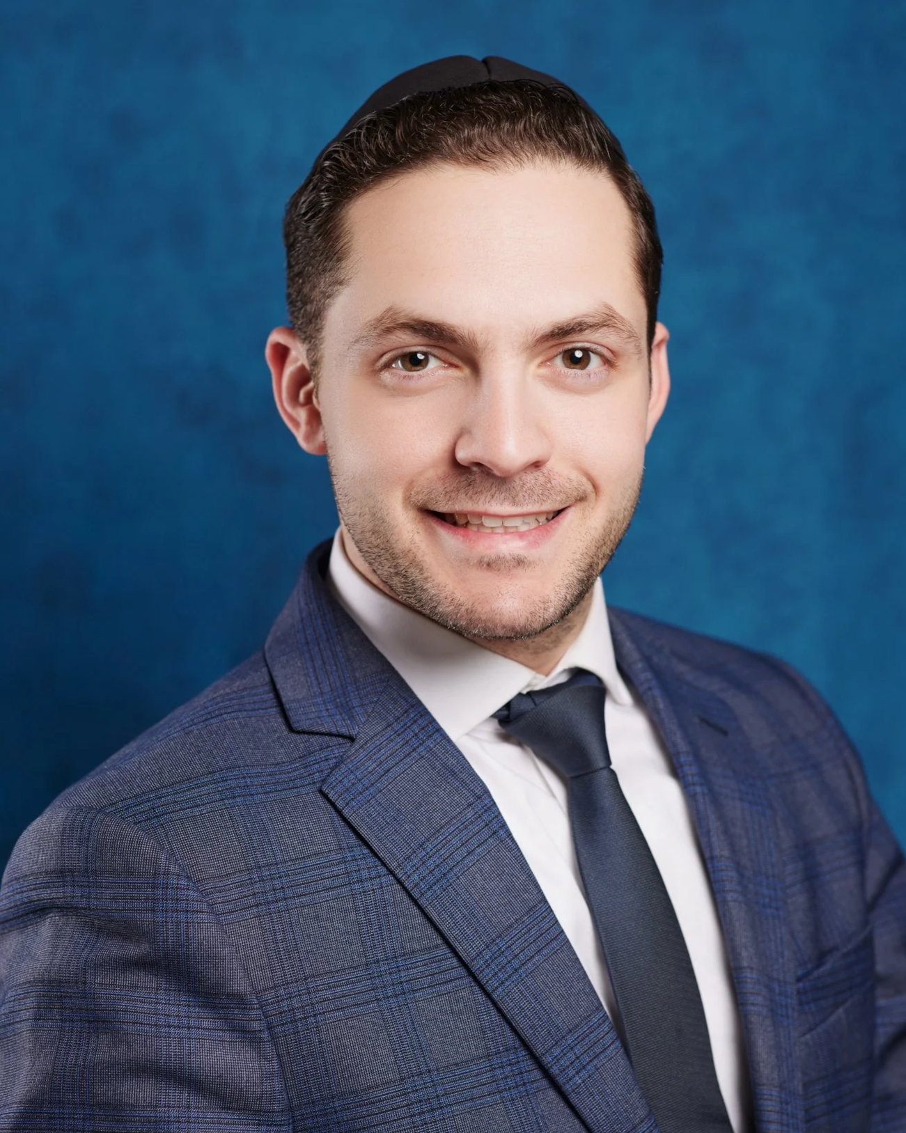Portrait of a young man in a checkered suit and tie, smiling, against a blue background.