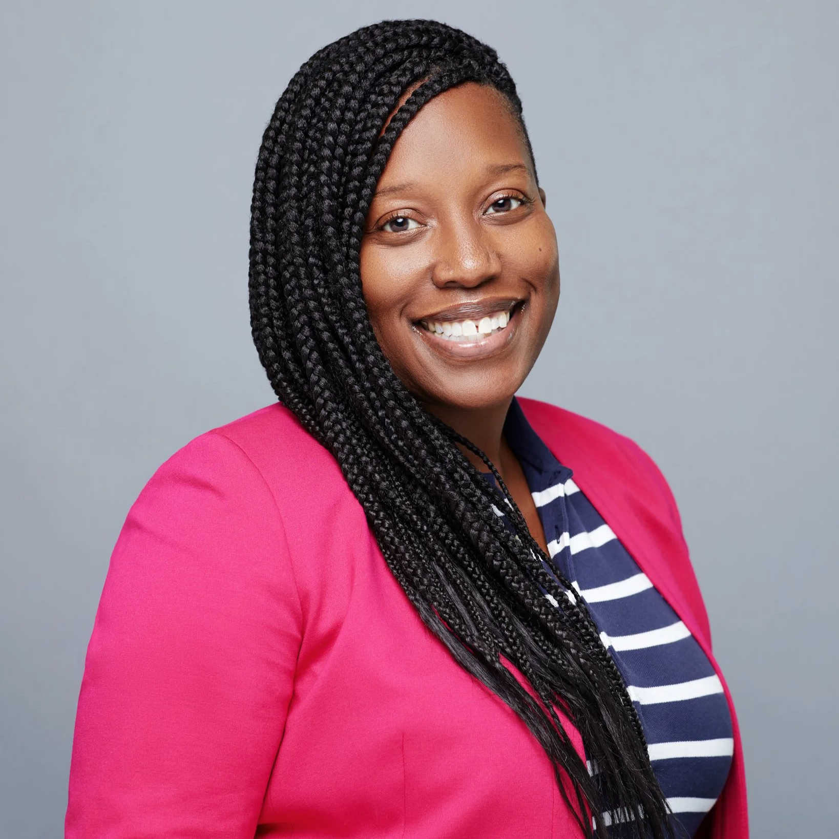 Portrait of a smiling woman with braided hair, wearing a pink blazer, striped shirt, and standing against a plain gray background.