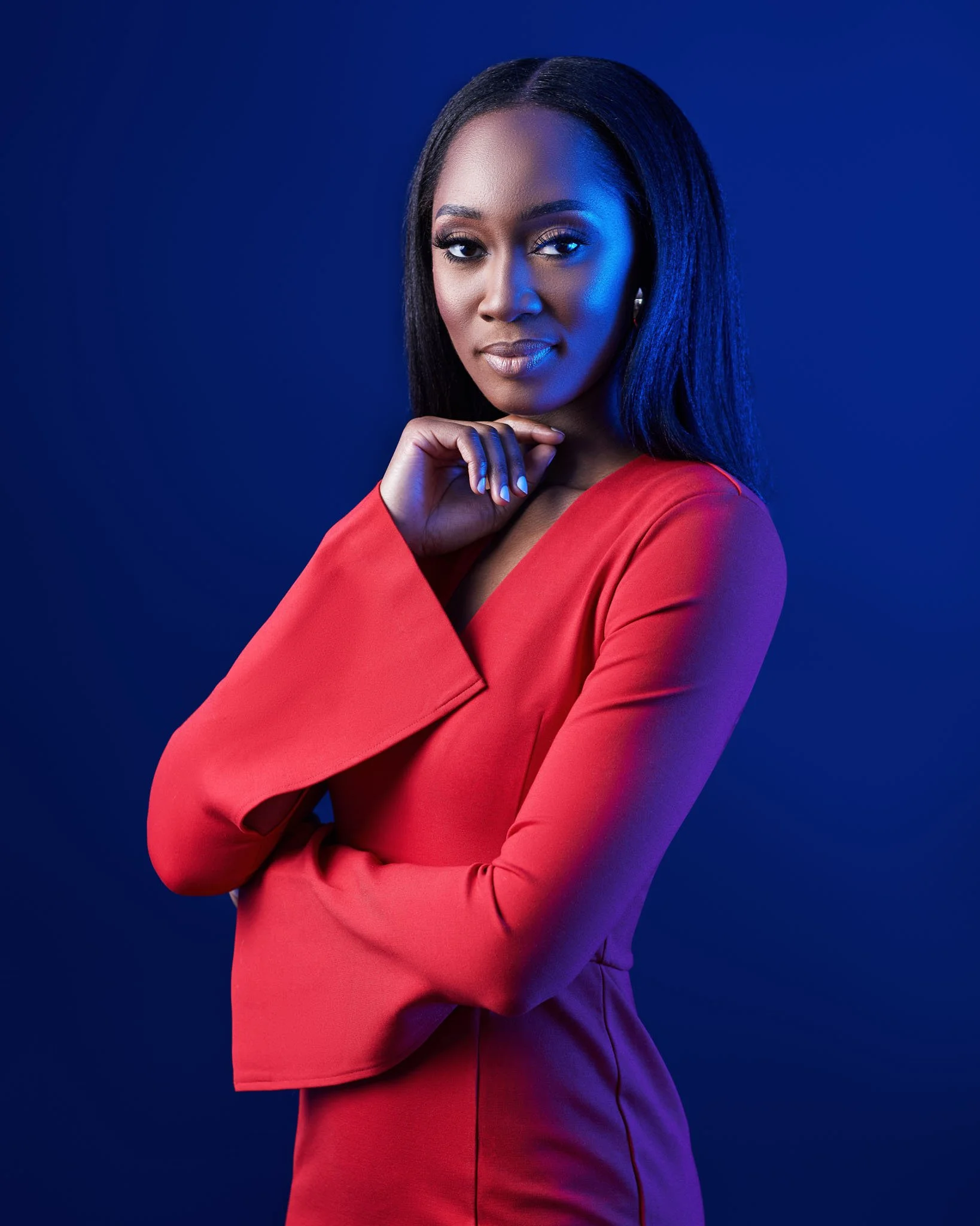 A woman with long straight black hair wearing a red top, posing with her hand near her chin against a dark blue background. She has a confident expression and is makeup with bold eye makeup.