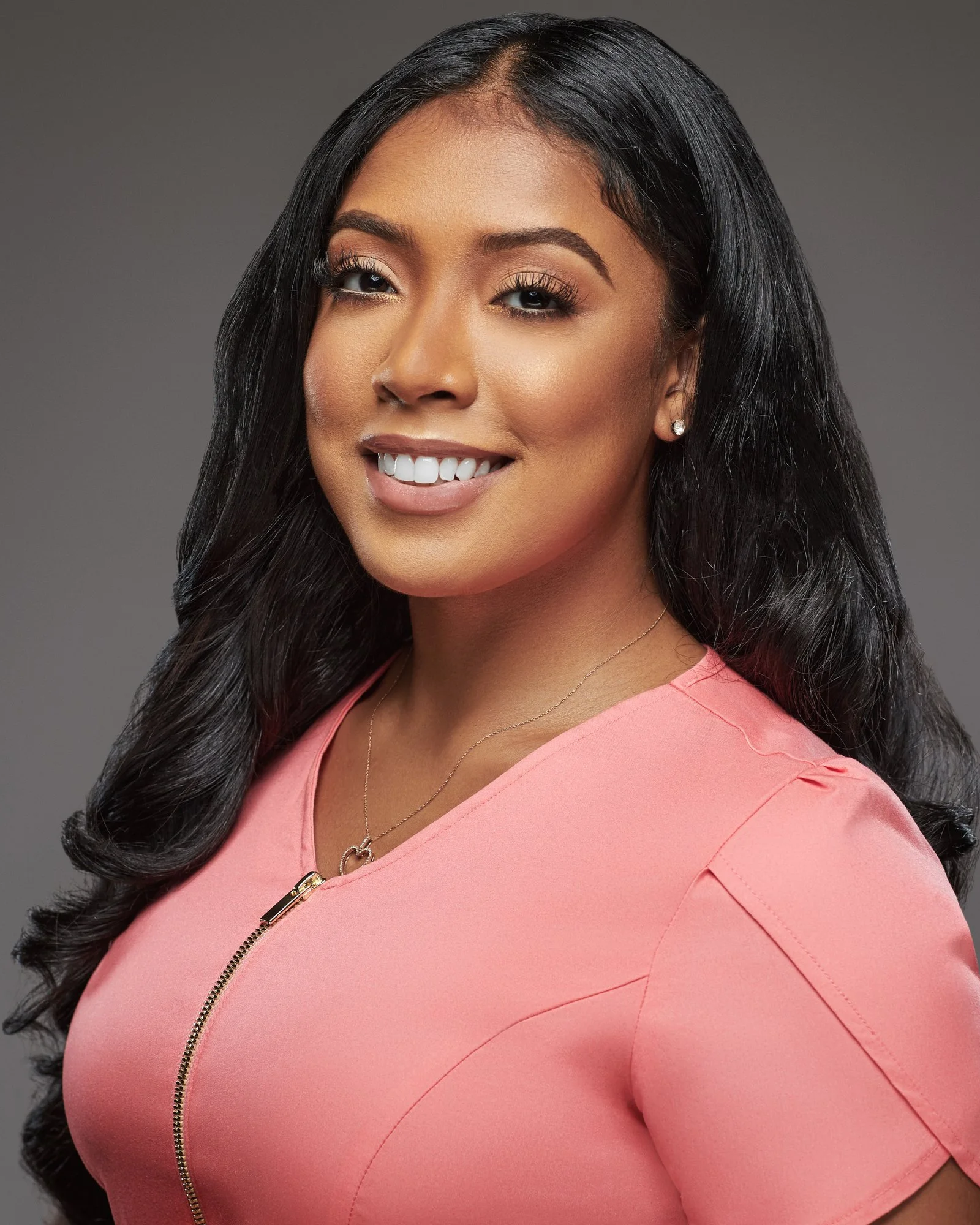 Close-up portrait of a smiling woman with long black hair, wearing a pink dress with a front zipper, jewelry, and earrings, against a neutral background.