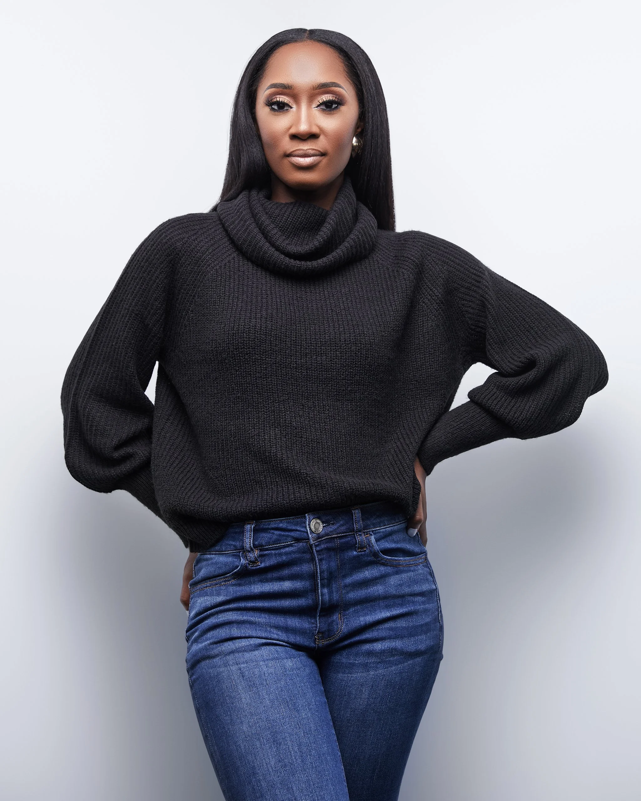 A confident woman with long black hair, wearing a black knit turtleneck sweater and blue jeans, standing against a plain white background.