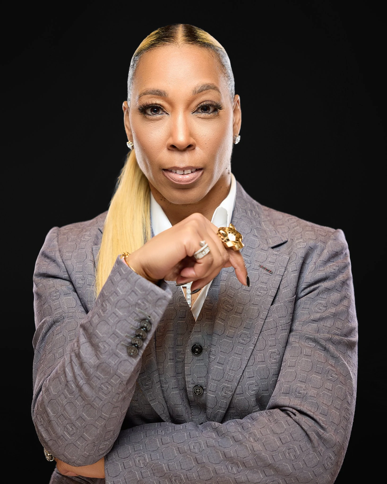 A woman with blonde hair slicked back, wearing a gray patterned blazer, white shirt, and various rings, earrings, and a bracelet, standing against a black background.