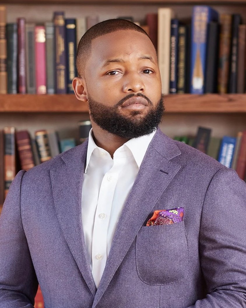A man with a beard wearing a white shirt and a gray suit jacket, standing in front of a bookshelf filled with books.