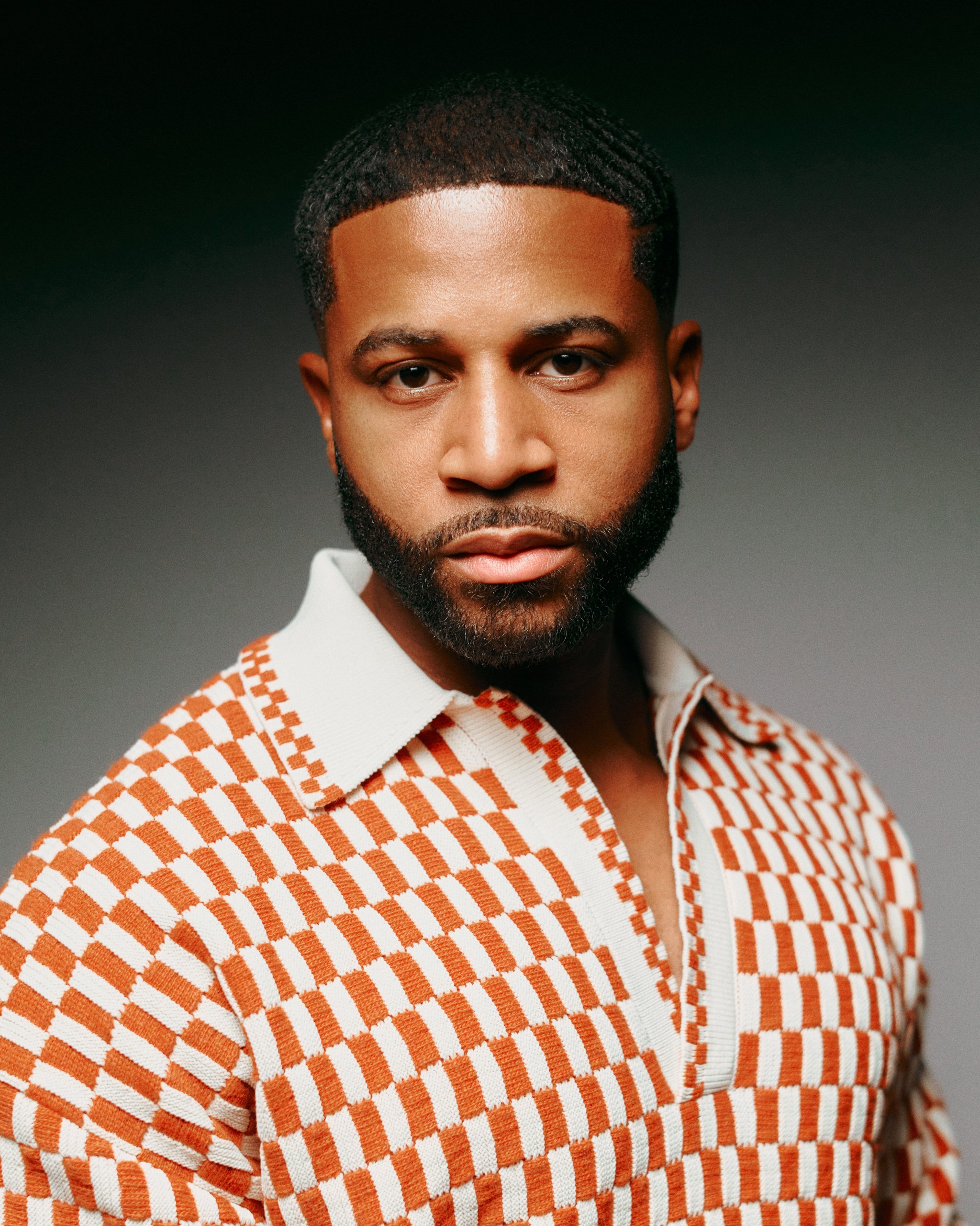 The image features a man with dark skin, a well-groomed beard, and styled hair, wearing a shirt with an orange and white checkered pattern. The background is dark and neutral, emphasizing his face and upper body.
