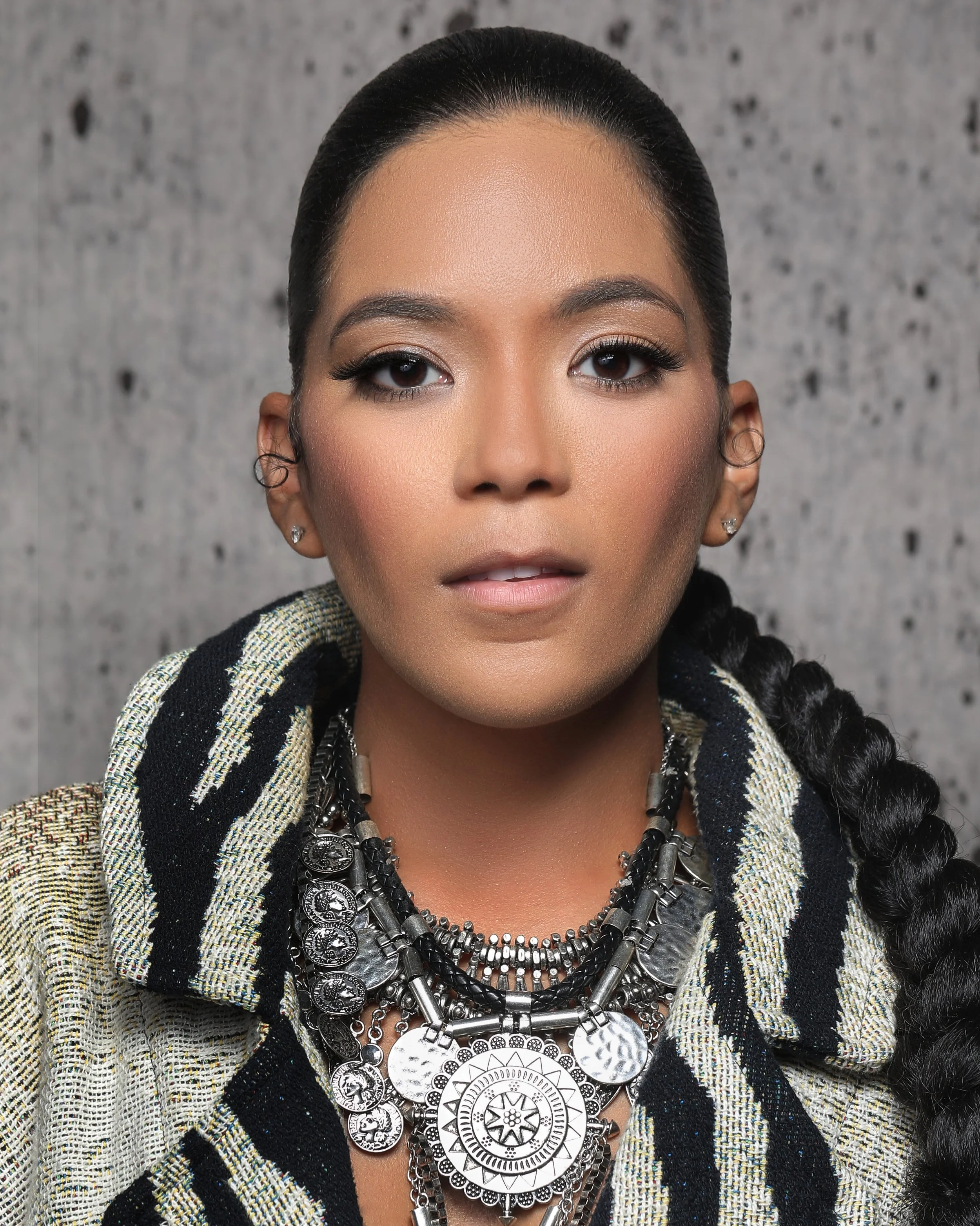 A woman with dark hair in a braid, wearing multiple layered silver necklaces, earrings, and a plaid blazer, standing against a textured gray background.