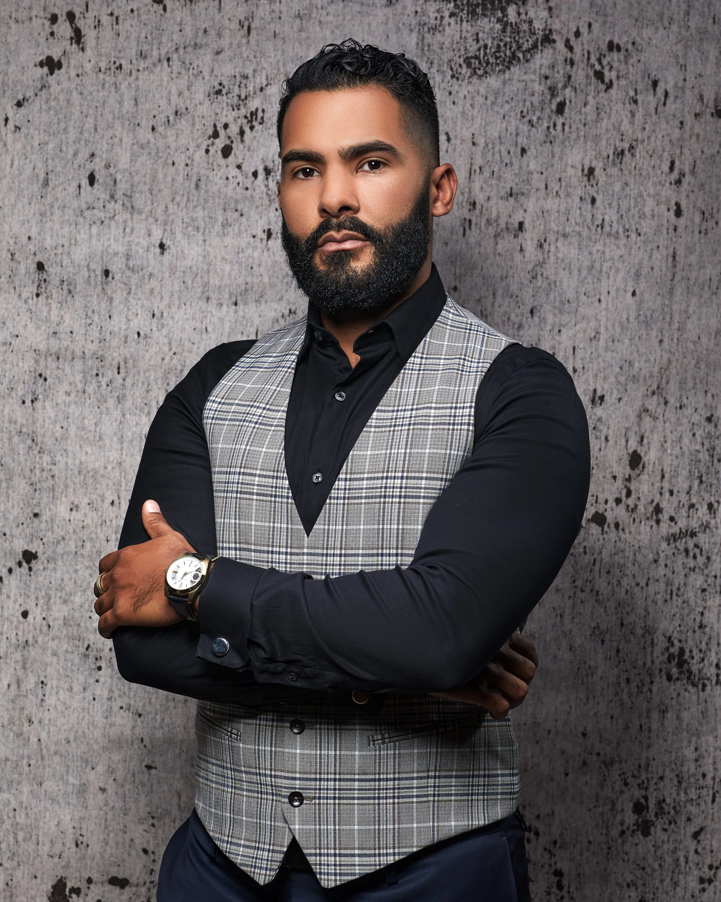 A man with a dark beard and short curly hair wearing a black shirt, checkered vest, and dark pants, standing with arms crossed in front of a textured wall.