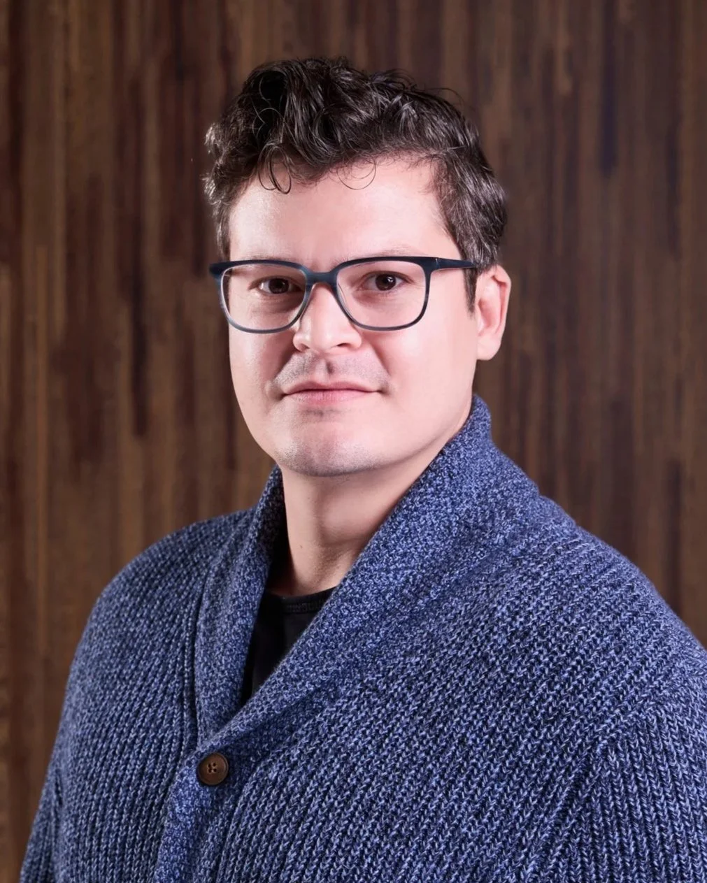 A portrait of a man with short dark curly hair, glasses, and a light skin tone, wearing a blue knit cardigan over a black shirt, standing against a wooden background.