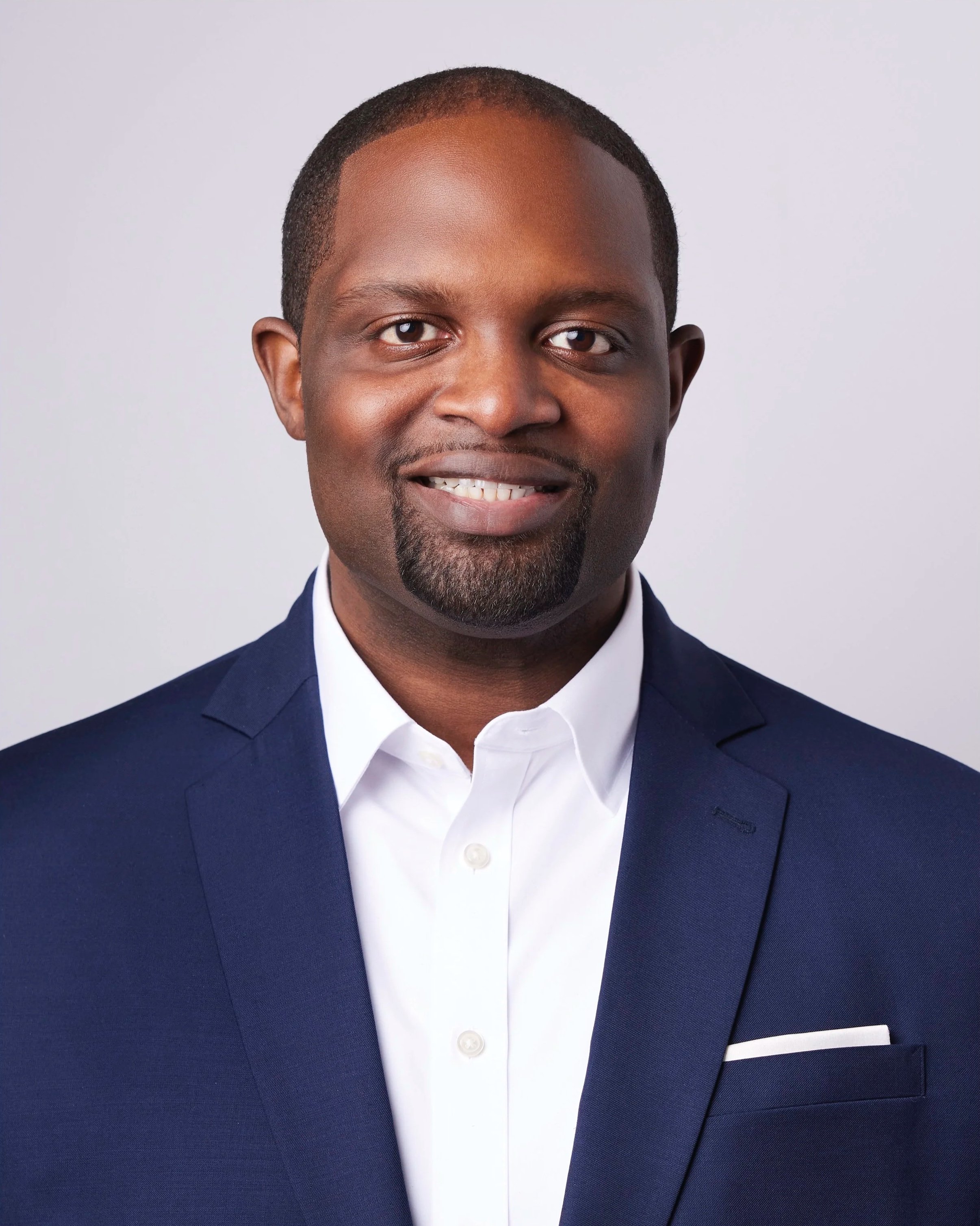 Close-up of a smiling man in a navy blue suit and white shirt against a light background.