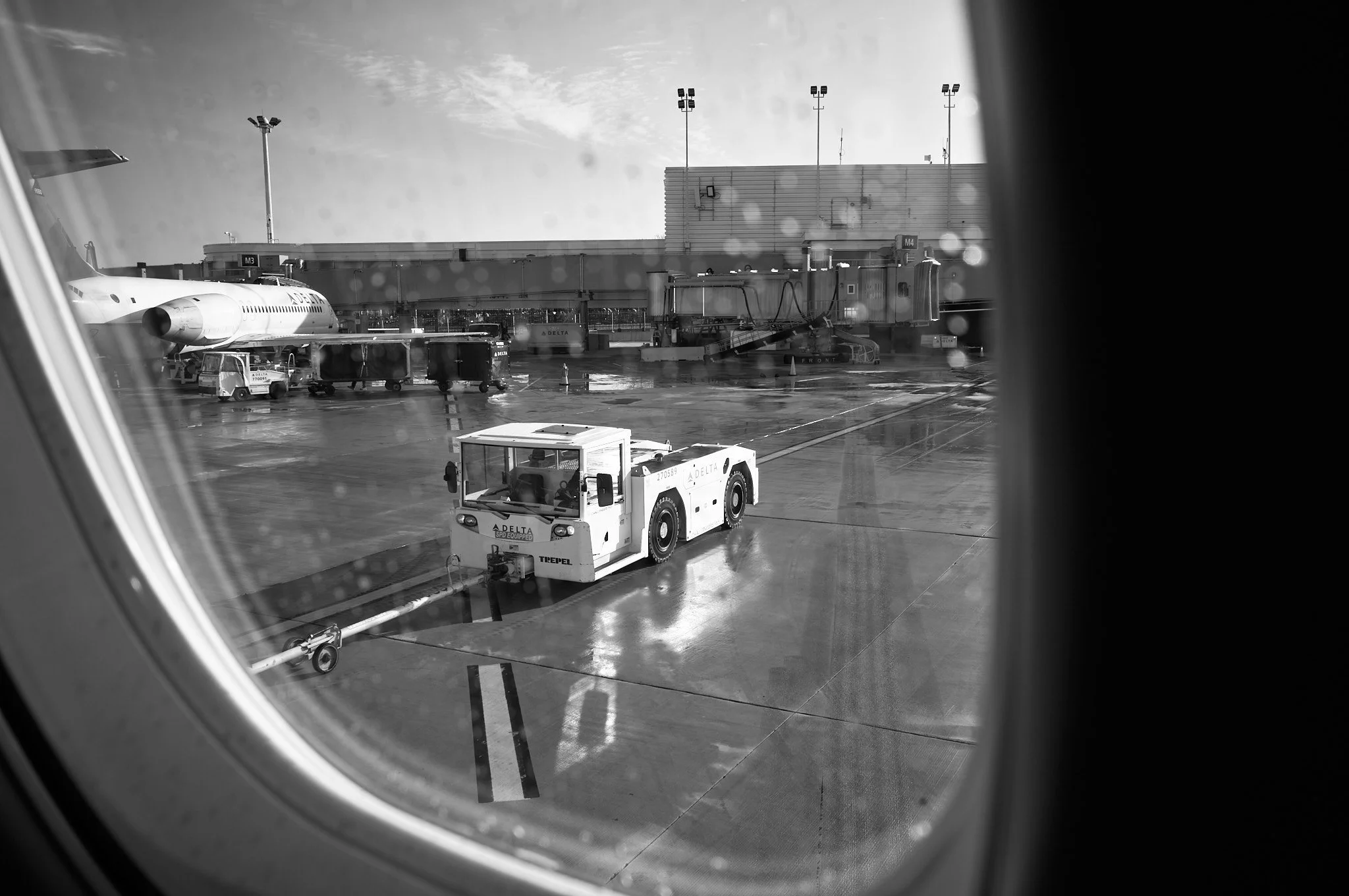 View of an airport tarmac seen through an airplane window, featuring a tug vehicle in the foreground, a Delta airplane, and airport structures and equipment in the background  by Matt Ellis