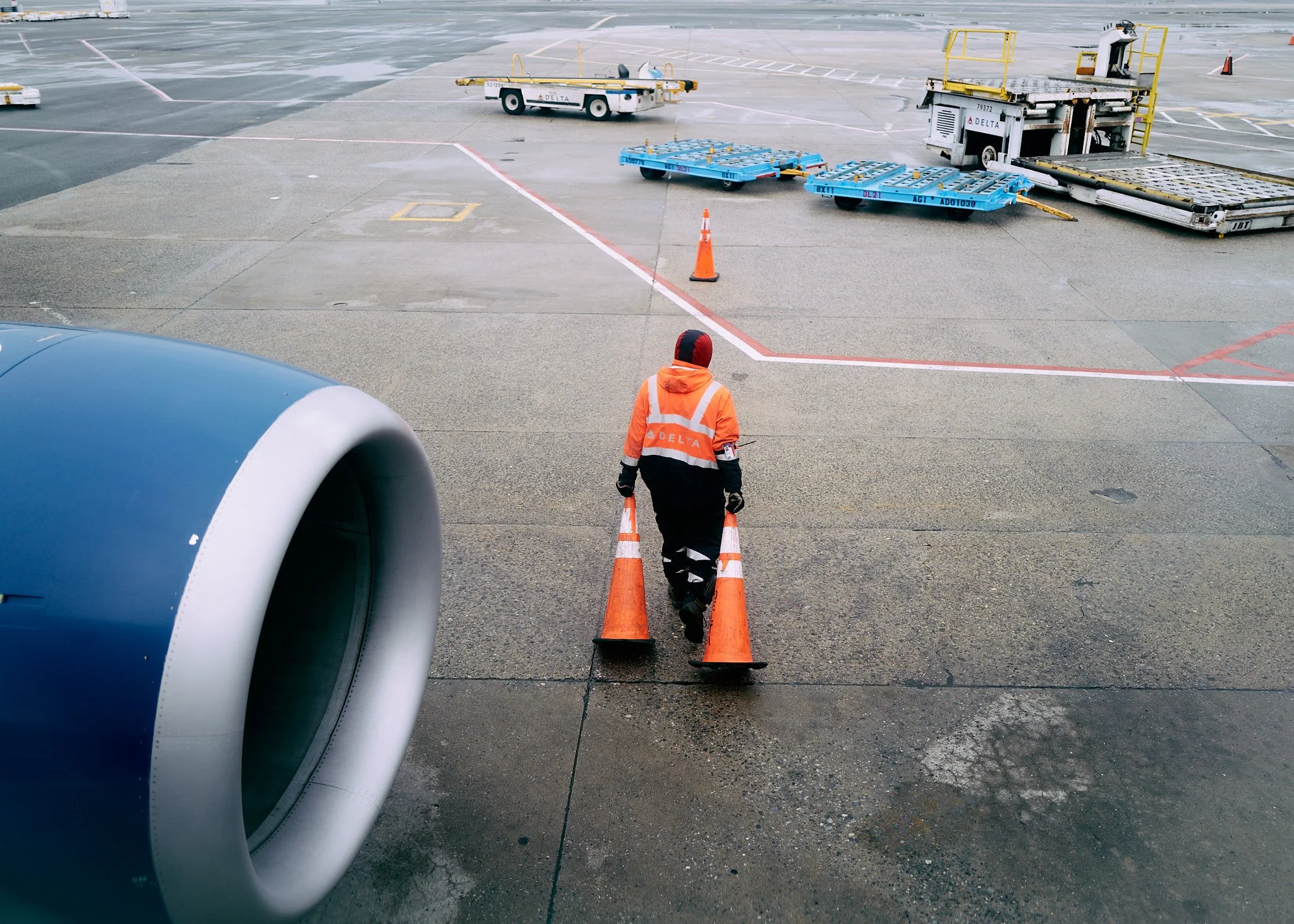A worker walking on an airport tarmac, pushing orange traffic cones behind him, near large airplane engine, with luggage carts and ground equipment visible in the background  by Matt Ellis