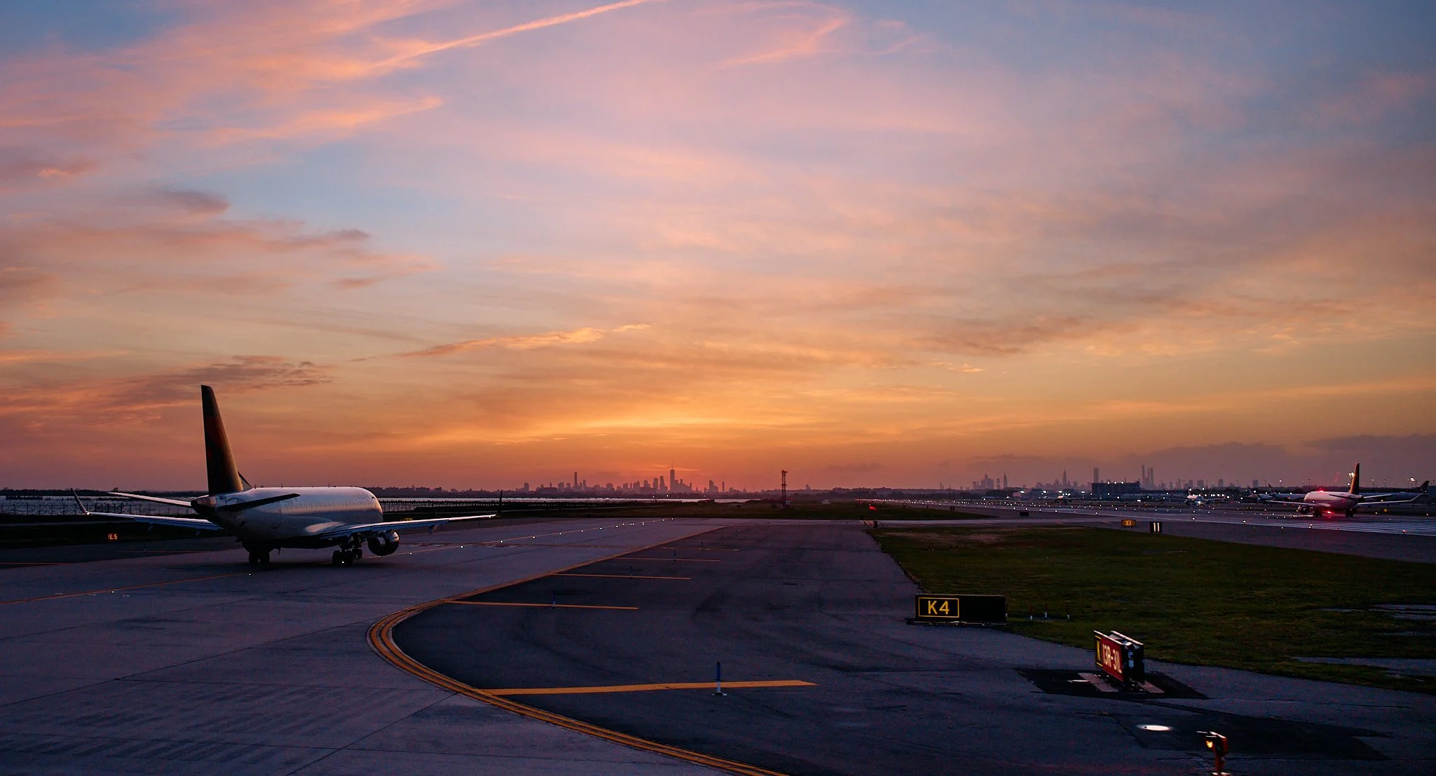 Jfk airport tarmac during sunset with airplanes parked, a city skyline in the distance, and colorful sky with pink and orange clouds.