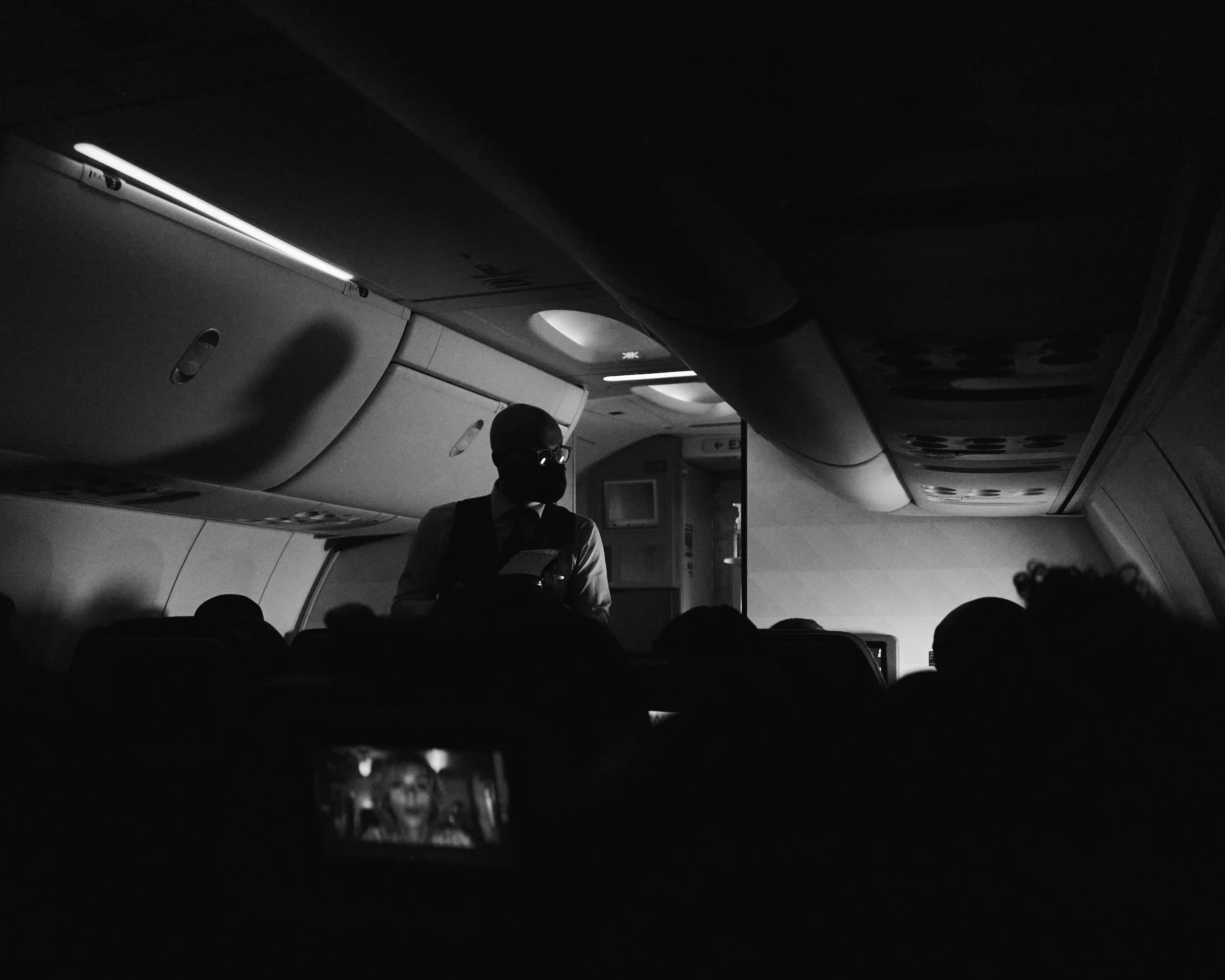 Black-and-white photo of a man with glasses and a face mask standing in the aisle of an airplane, illuminated by overhead lights, with passengers seated around him.