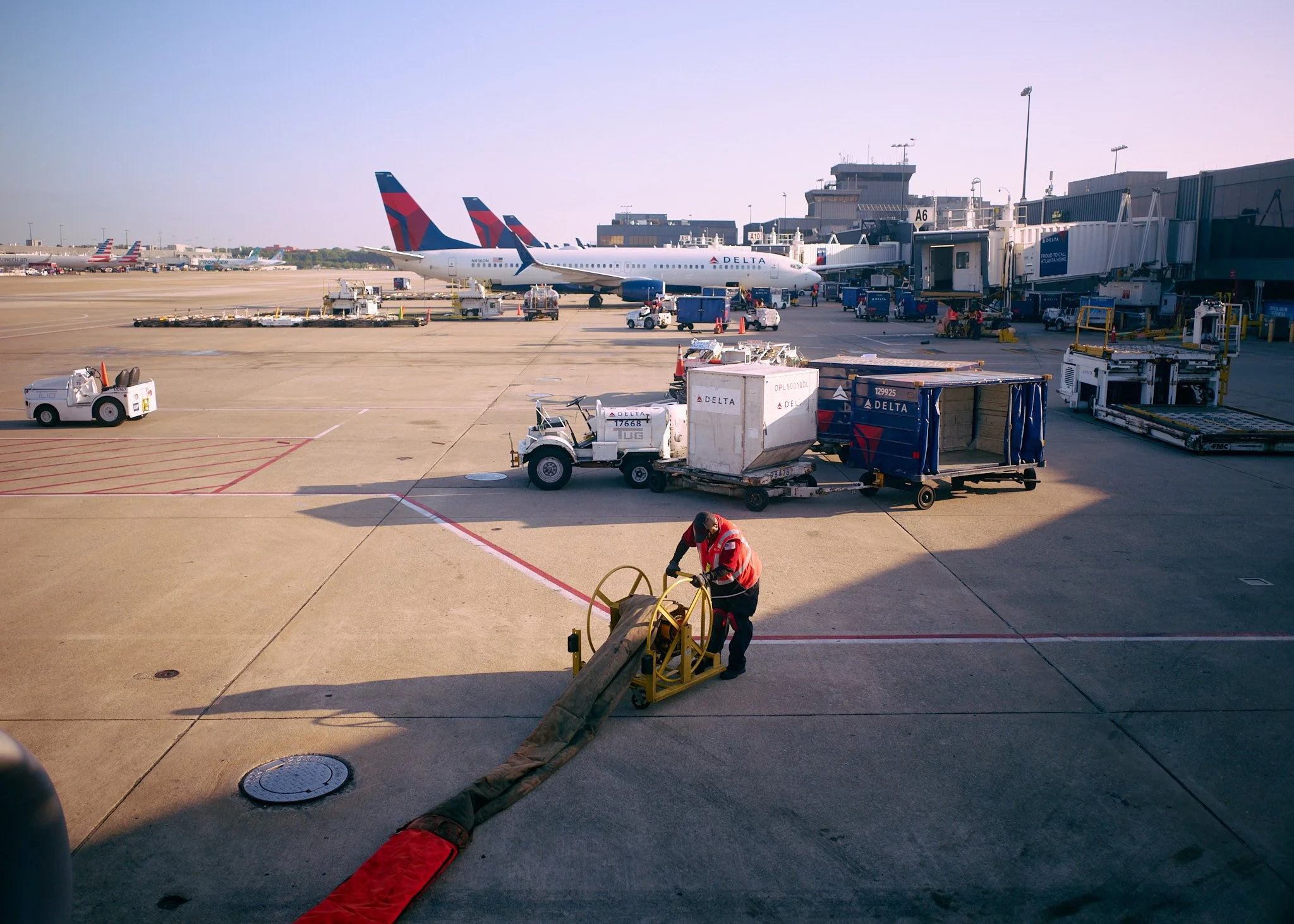 Airport tarmac with Delta airplanes parked at gates, ground crew member adjusting a hose, various baggage carts, and ground support equipment  by Matt Ellis
