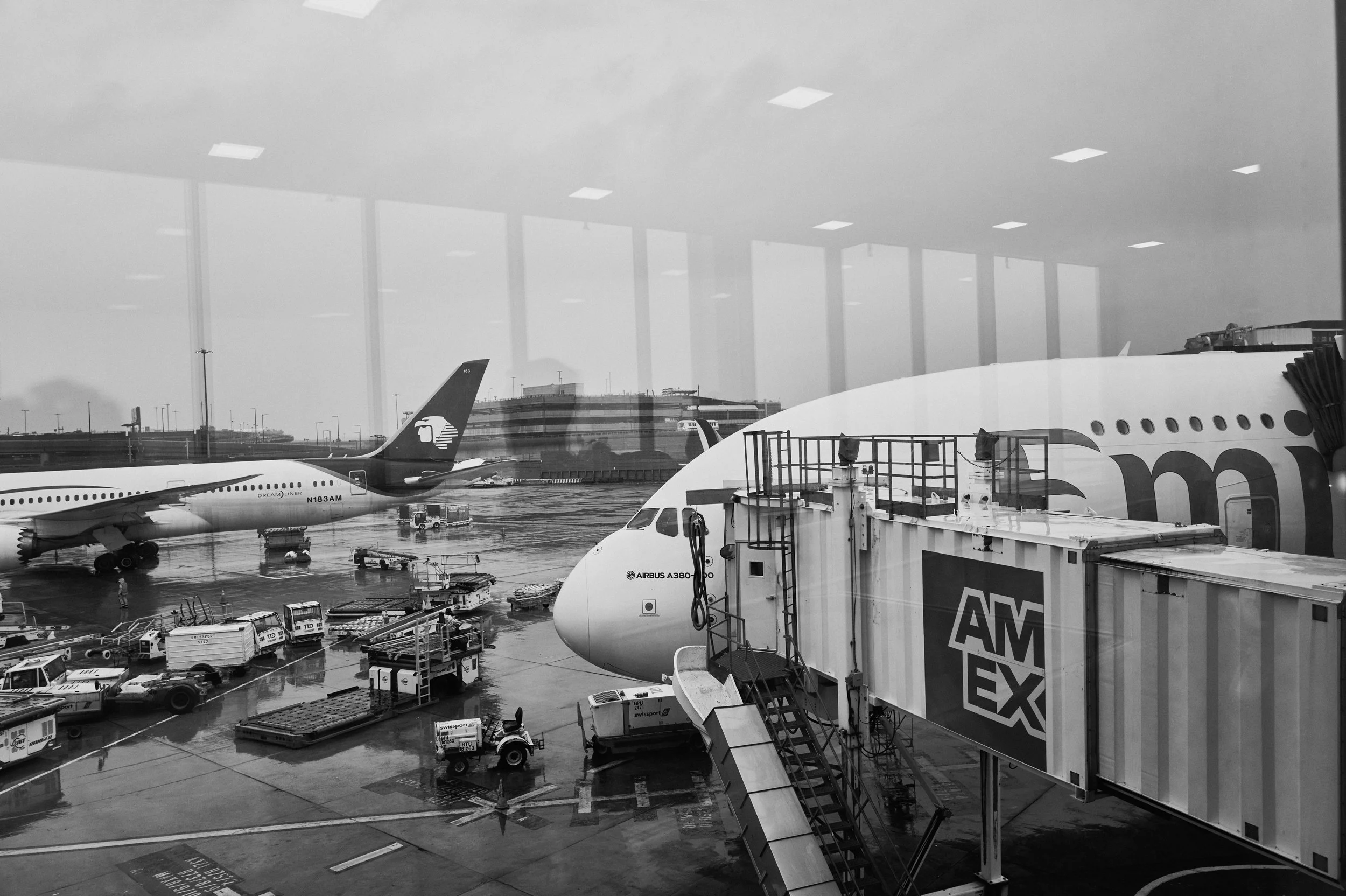 View of an airport terminal through a window, showing airplanes parked at gates, baggage carts, airport vehicles, and a cloudy sky outside.
