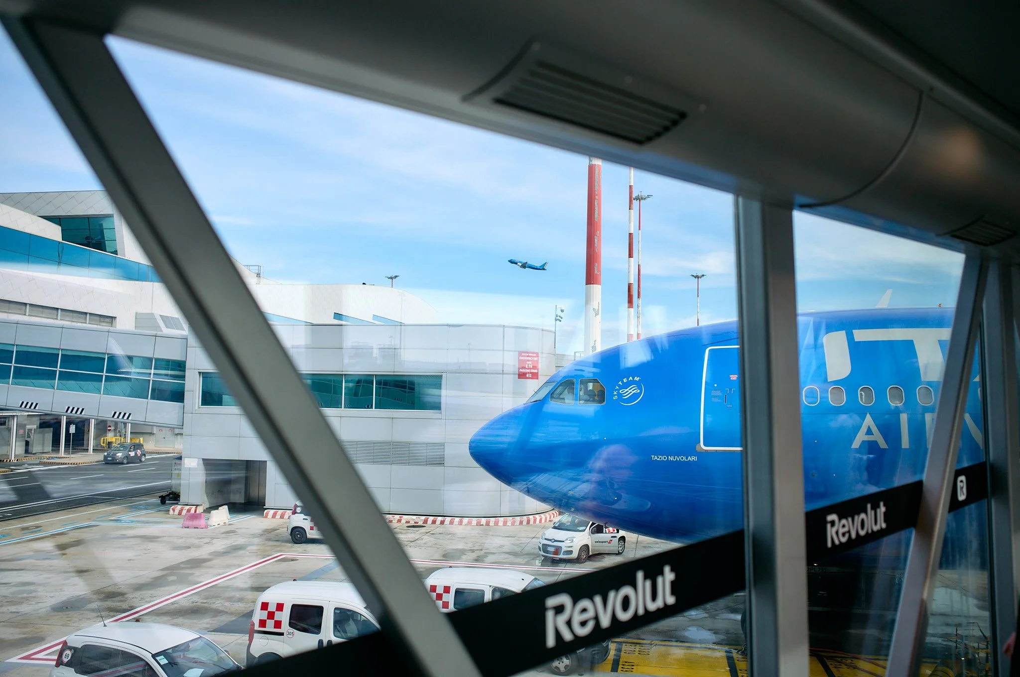 View of an airplane parked at the airport gate with a jet bridge connecting to the plane, window view showing another airplane taking off in the sky, and airport vehicles on the tarmac  by Matt Ellis