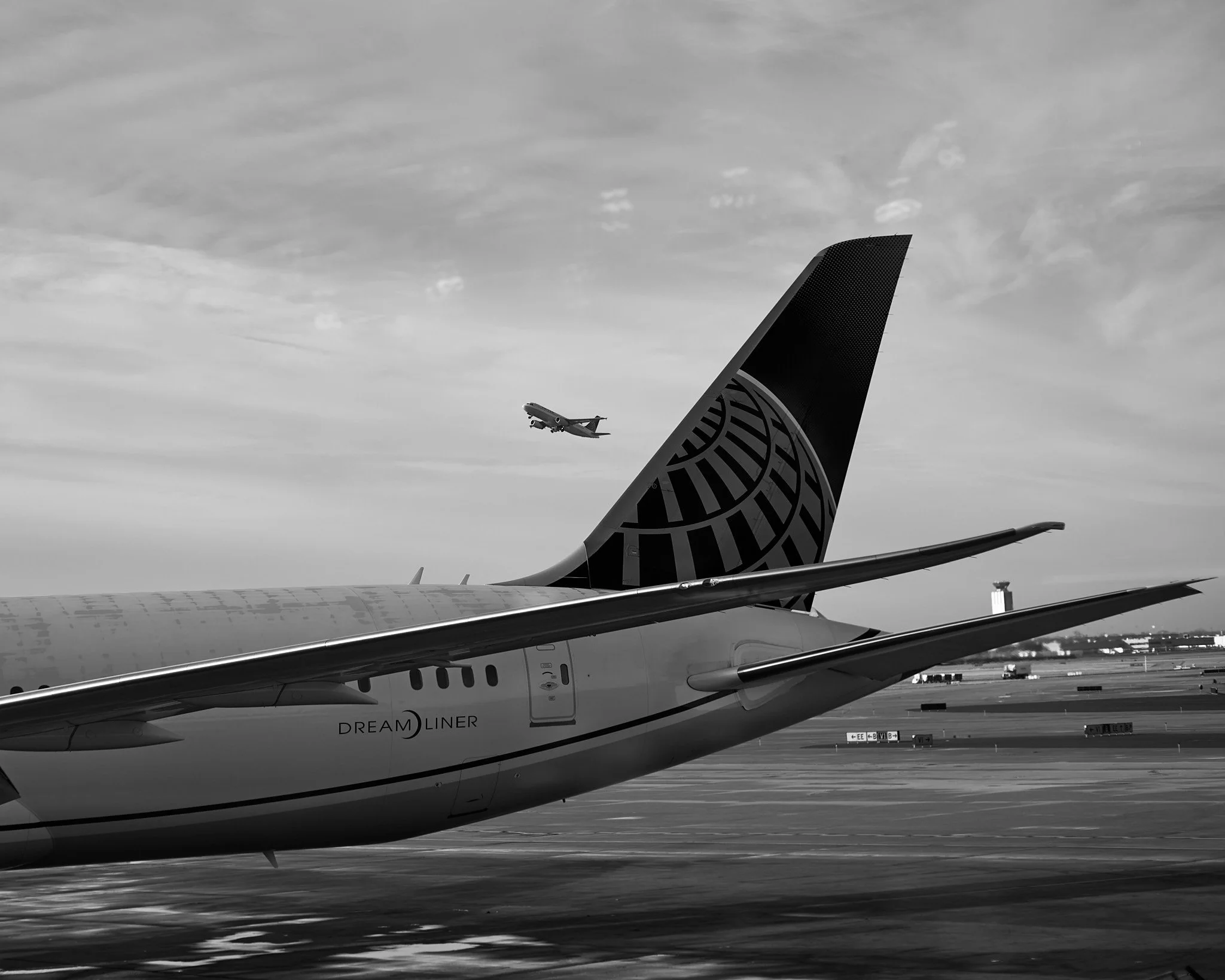 A close-up of a commercial airplane on the tarmac with its tail and wing visible, and another airplane flying in the sky in the background, with some clouds overhead  by Matt Ellis