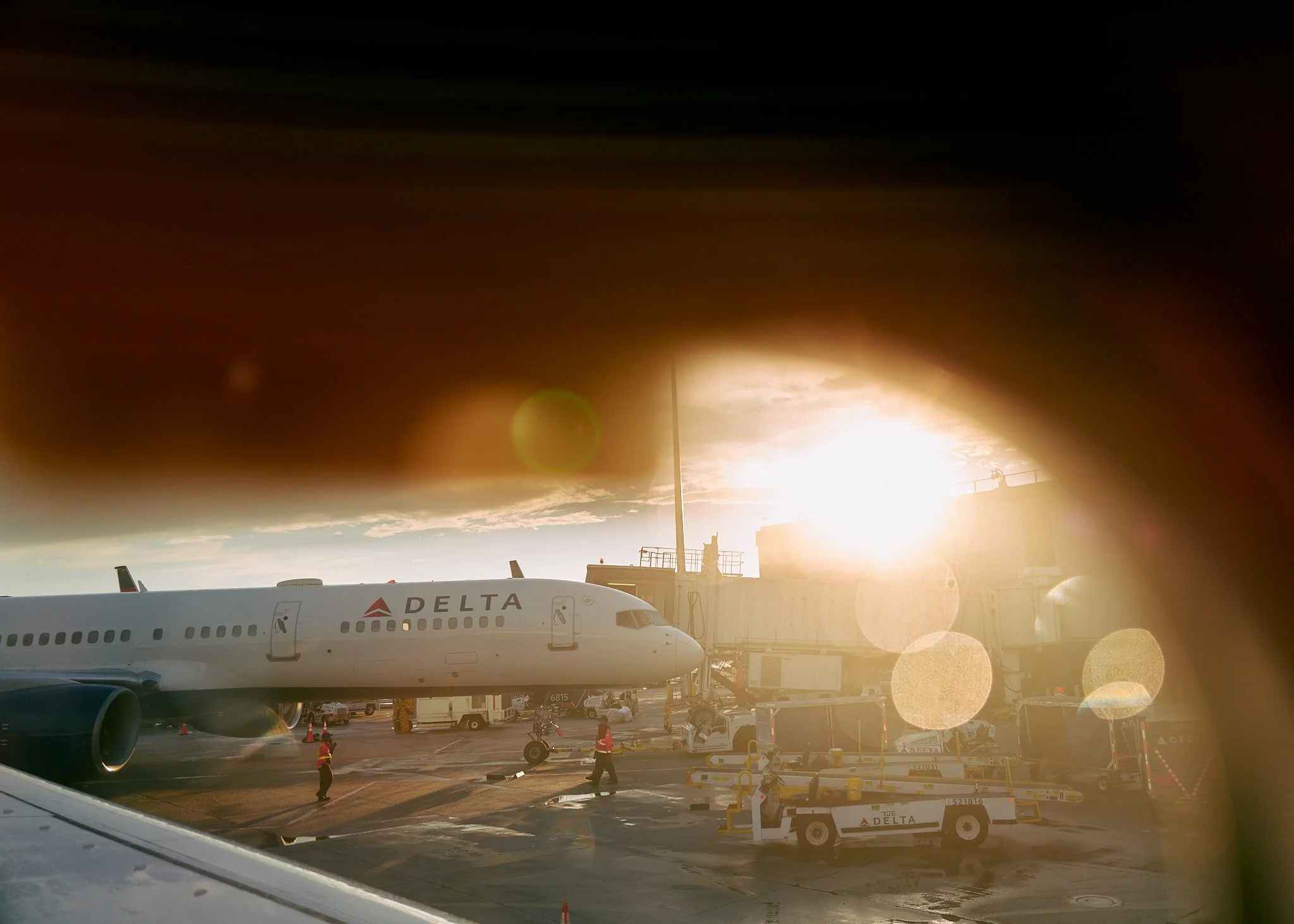 An airport tarmac with a Delta airplane parked beside a boarding bridge, ground crew working, and the sun setting in the background  by Matt Ellis