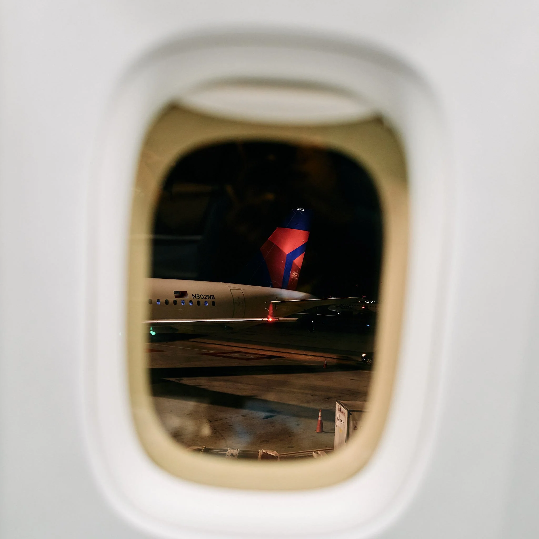 View through an airplane window at night showing a Delta airplane on the tarmac with part of its tail visible with the airline's logo.