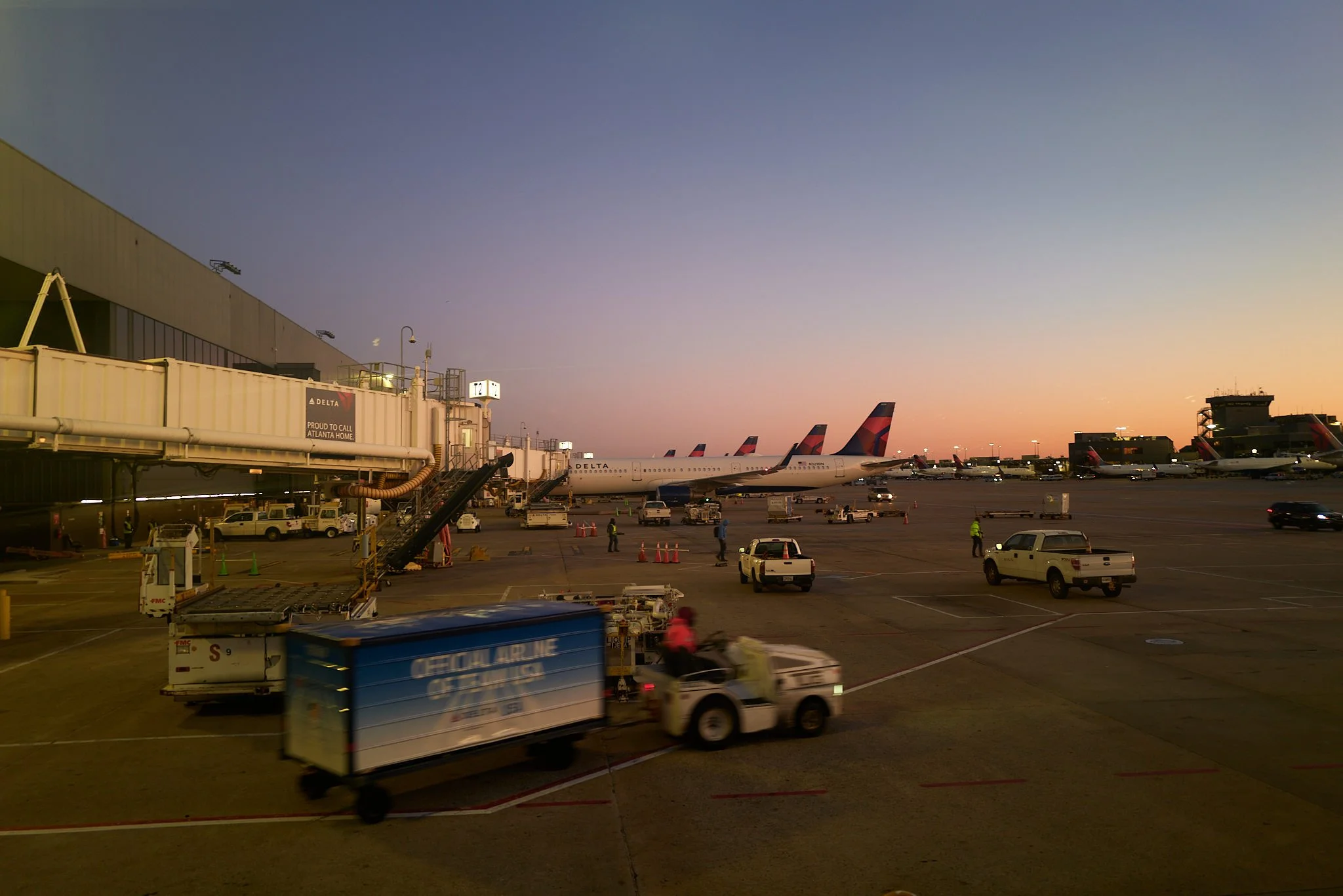 Airport tarmac at sunset with several Delta airplanes parked, ground crew working, airport vehicles moving, and a sky transitioning from blue to orange  by Matt Ellis