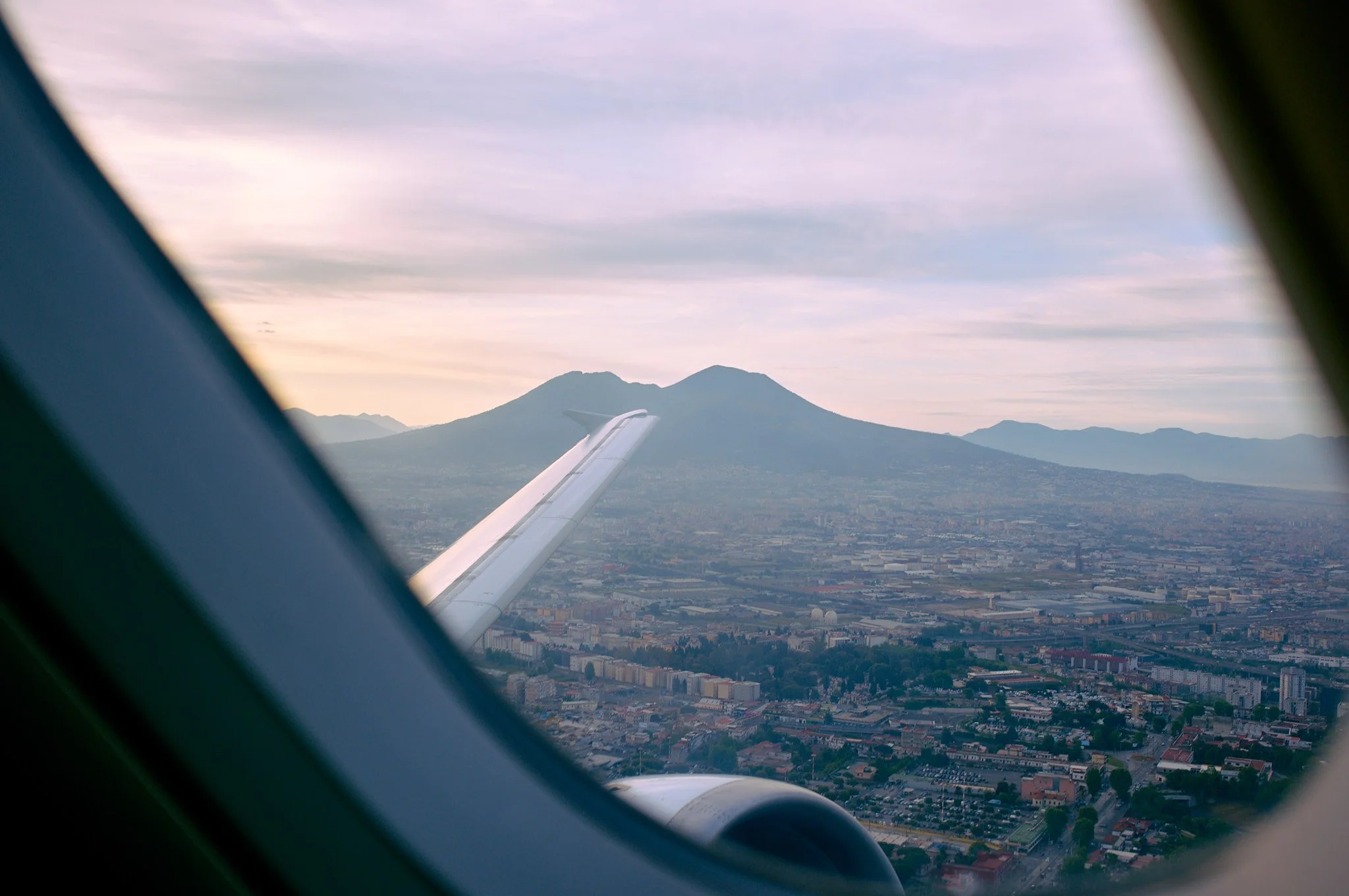 View of a volcano in Guatemala from an airplane window with the airplane wing visible in the foreground, cityscape below, and cloudy sky.