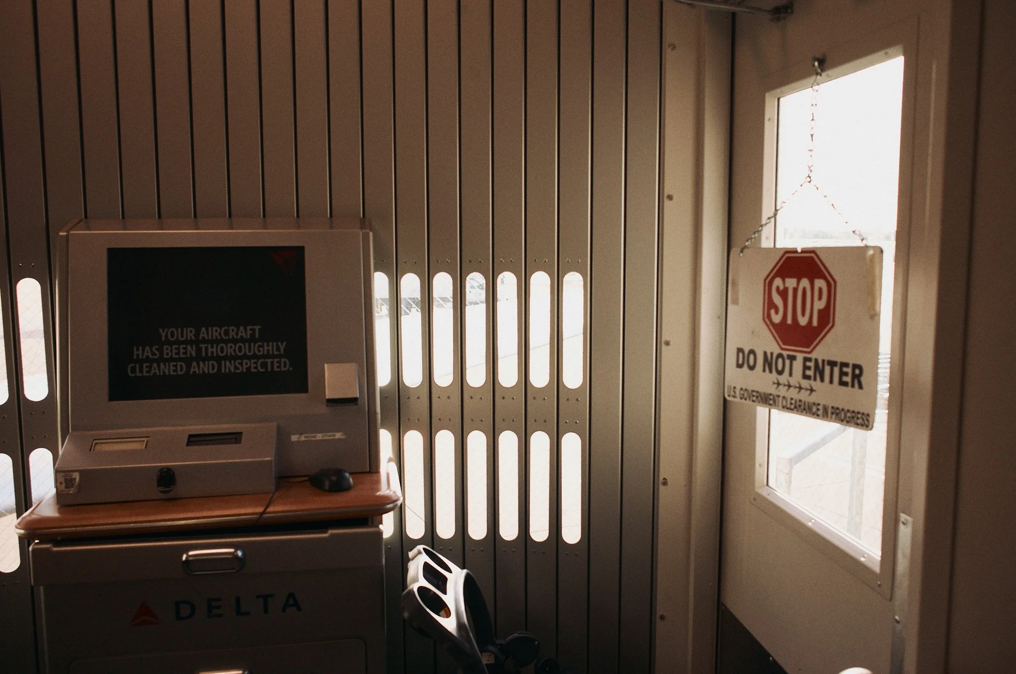 An airport security area with a computer screen displaying a message about aircraft cleaning. There is a small luggage scanner beneath the screen. A large metal door with vertical slots and a window with a 'STOP DO NOT ENTER' sign is on the right sid